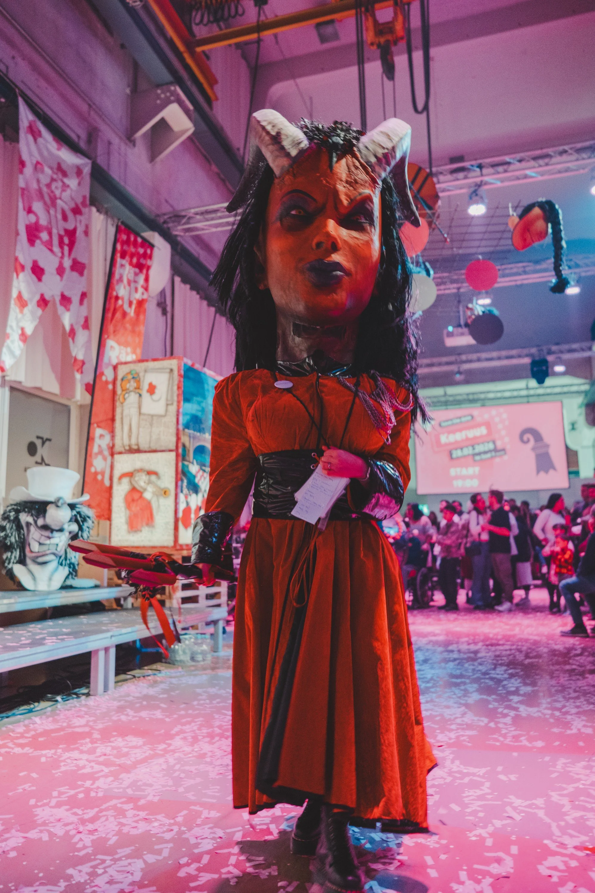 Großes Teufelsmaske mit Hörnern und schwarzen Haaren, getragen von einer Person in rotem Kleid bei einer Kostümparty oder Veranstaltung.