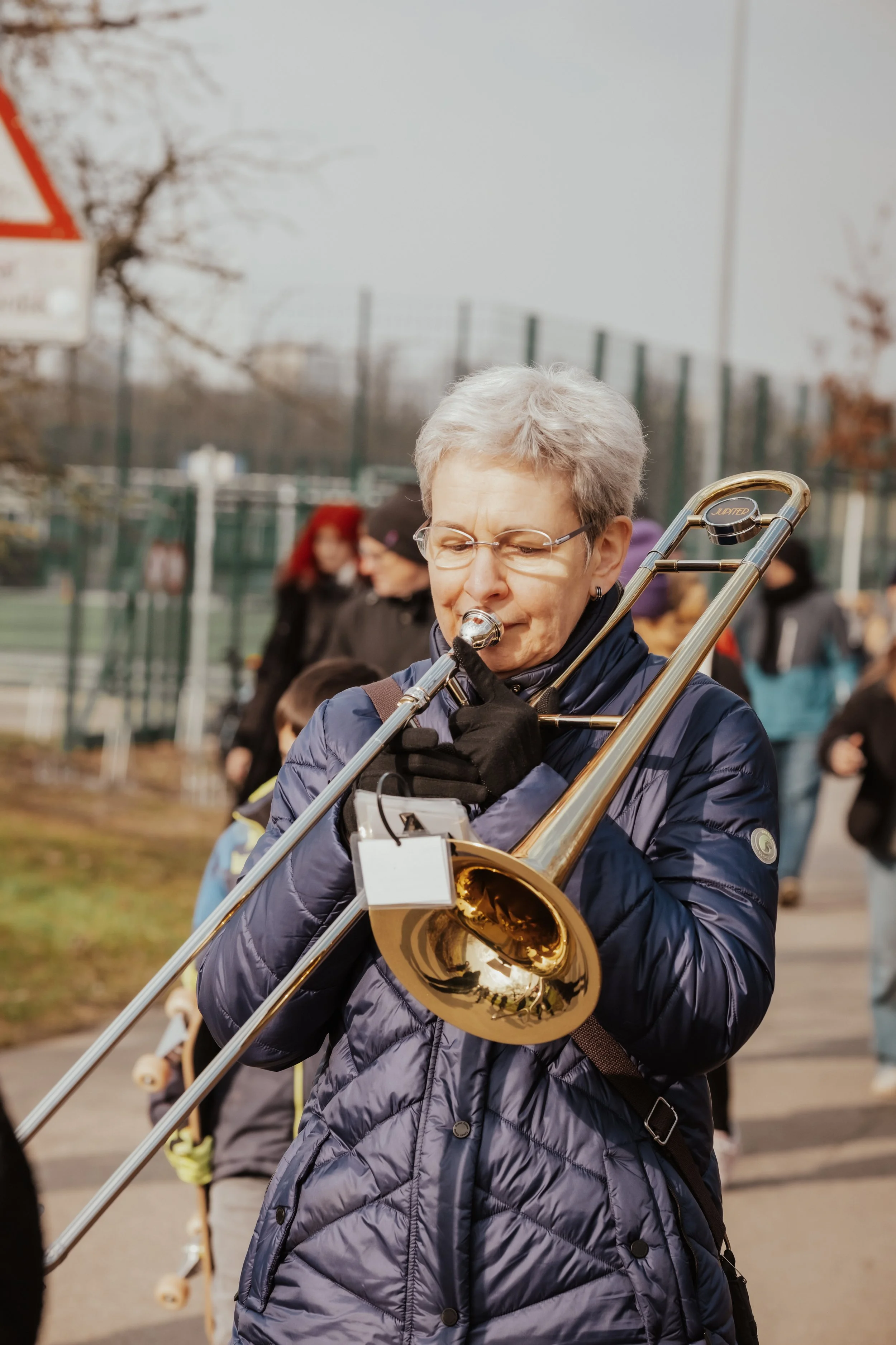 Eine ältere Frau spielt Trompete im Freien, umgeben von anderen Menschen und einem Tennisplatz im Hintergrund.