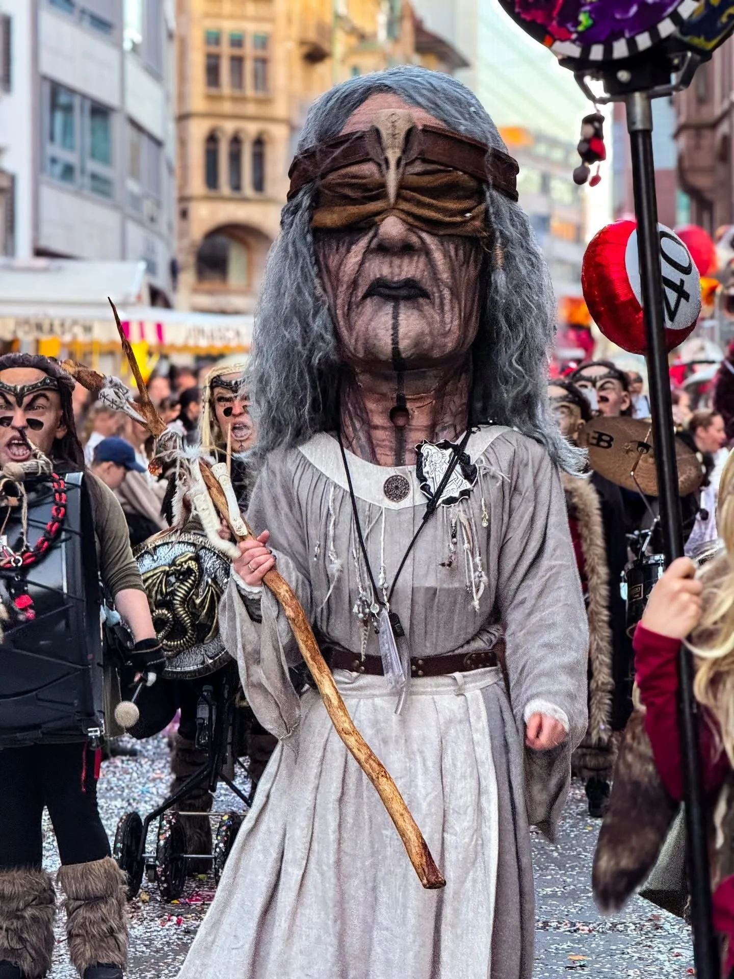 Person in Schildkrötkostüm bei Karnevalsfeier, umgeben von anderen Festteilnehmern in Kostümen, auf einer Straße mit confetti auf dem Boden.