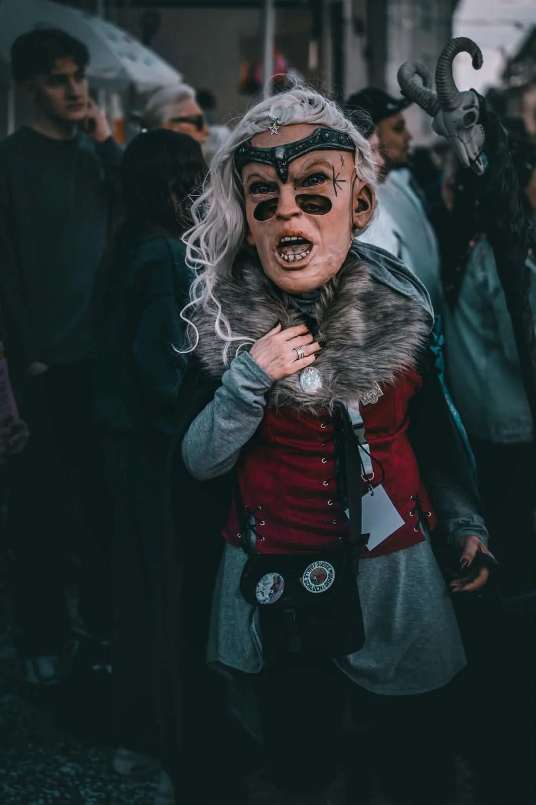 Person in Kostüm mit Maskerade und dekorativer Maske, umgeben von Menschen bei einer öffentlichen Veranstaltung oder Feier in einer Stadt.