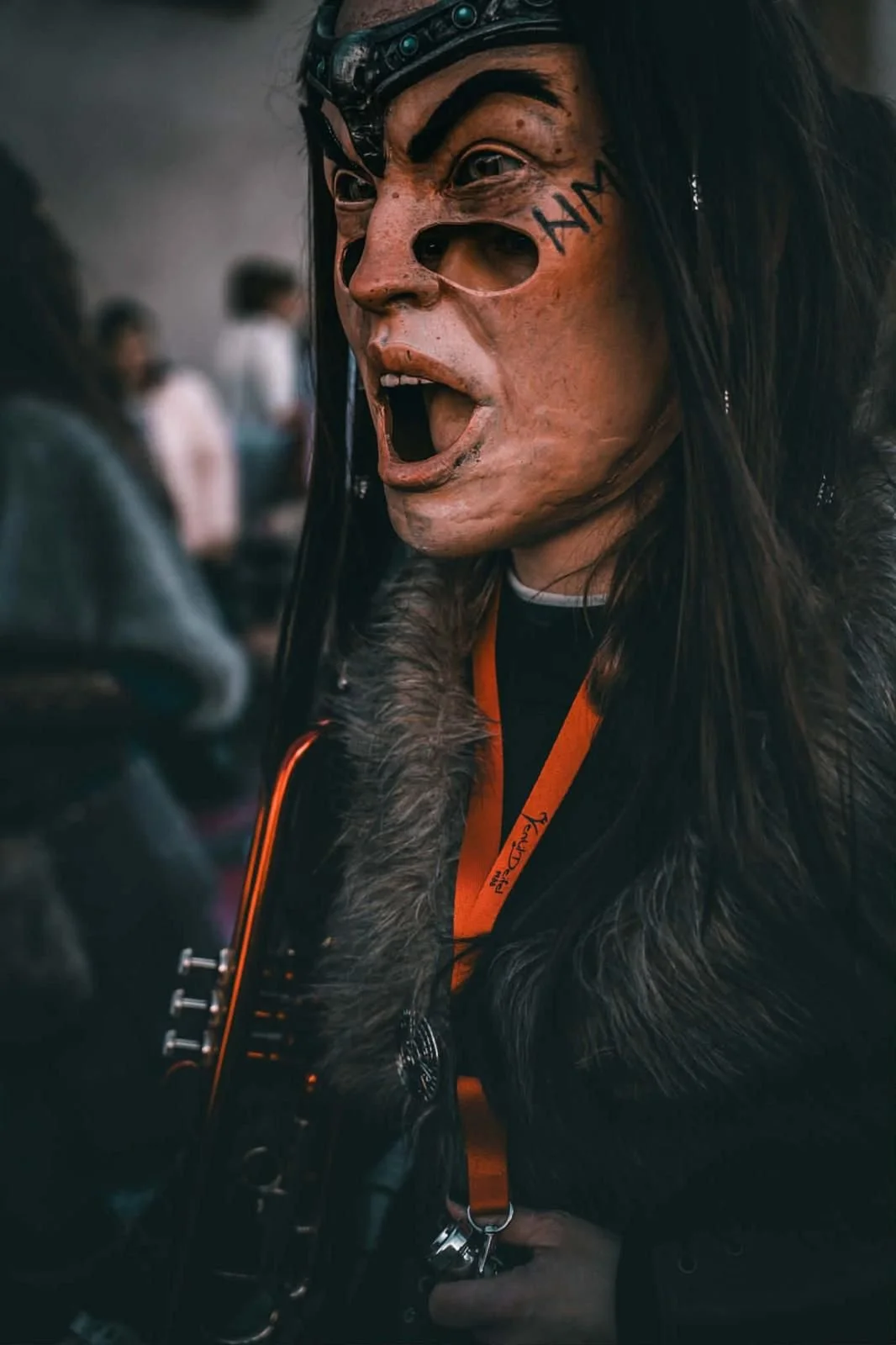 Eine Person trägt eine detaillierte Maske im wilden Fantasy- oder Horror-Stil mit Markierungen auf dem Gesicht, eine Lederjacke mit Pelzbesatz und eine Orange-Schnur um den Hals. Sie hält ein Musikinstrument, vermutlich eine Gitarre.