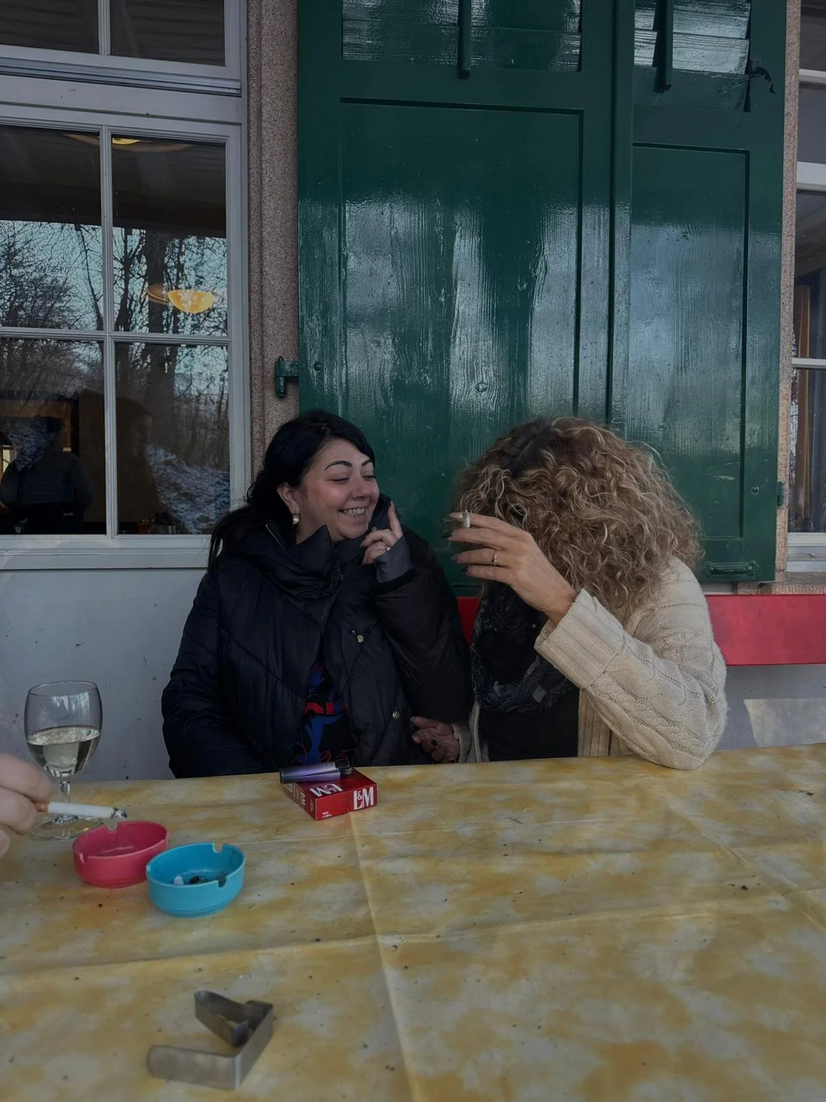 Zwei Frauen sitzen an einem Tisch, lachen und sprechen miteinander, während sie Zigaretten rauchen.