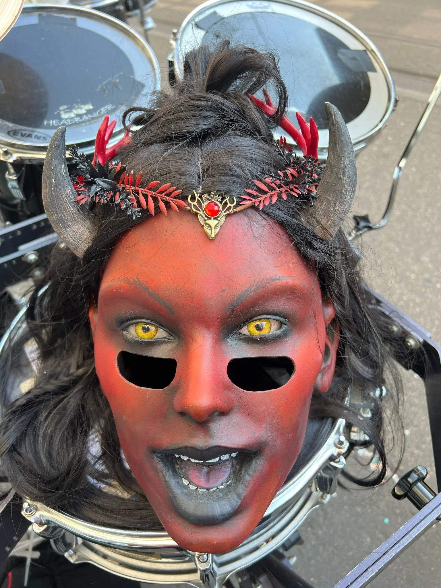 Eine Person trägt eine dämonisch aussehende Maske mit roten, schwarzen und schwarzen Akzenten, gelben Augen, schwarzen Lippen, und Hörnern. Sie trägt einen Kopfkranz mit roten und schwarzen Blättern und einer kleinen goldenen Dekoration mit rotem Ede