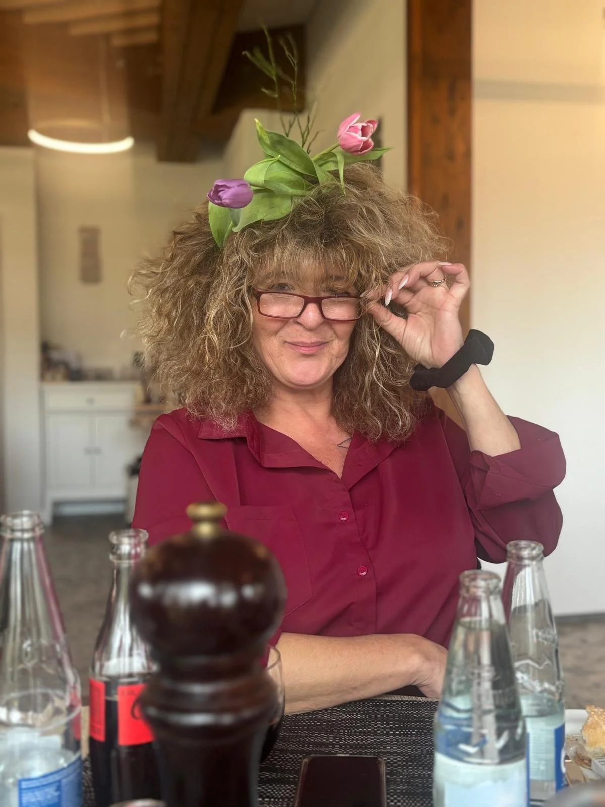 Frau mit Brille trägt einen roten Kleid und hat einen Blumenkranz mit Tulpen auf dem Kopf. Sie sitzt an einem Tisch mit Flaschen und hat eine entspannte Haltung.