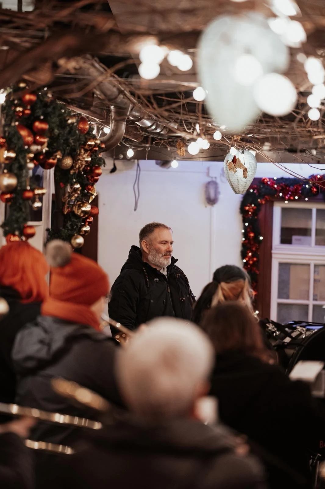 Eine Gruppe von Menschen, die in einem festlich dekorierten Raum sitzen, während ein Mann steht und spricht. Die Dekoration umfasst Weihnachtskugeln und Lichter.