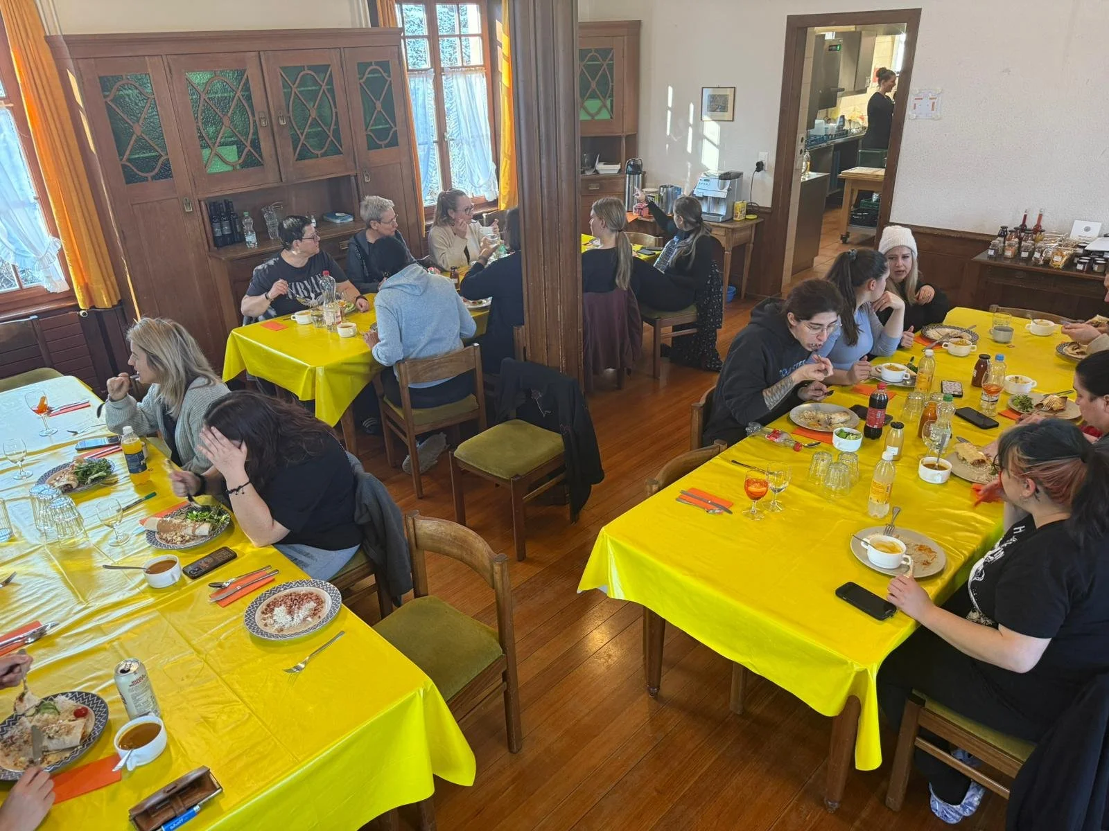 Mehrere Menschen sitzen an Tischen mit gelben Tischdecken in einem Raum und essen. Im Hintergrund ist eine Küche sichtbar.