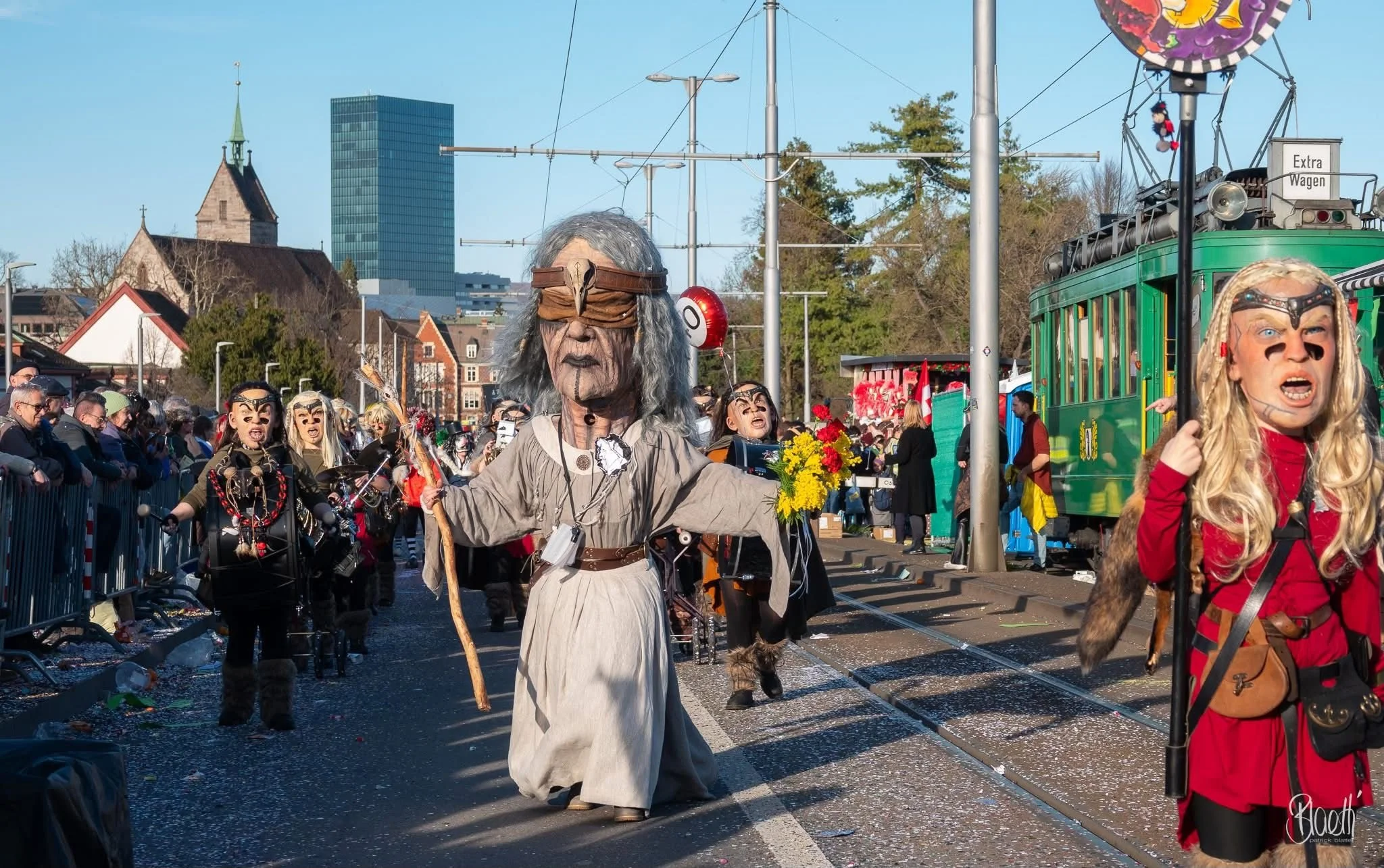 Menschen in Karnevalskostümen bei einer Parade, einige tragen riesige Masken und Kostüme, andere sind in bunte Kleidung und Accessoires gekleidet, im Hintergrund sind Zuschauer, Straßenbahn und Stadtgebäude sichtbar.