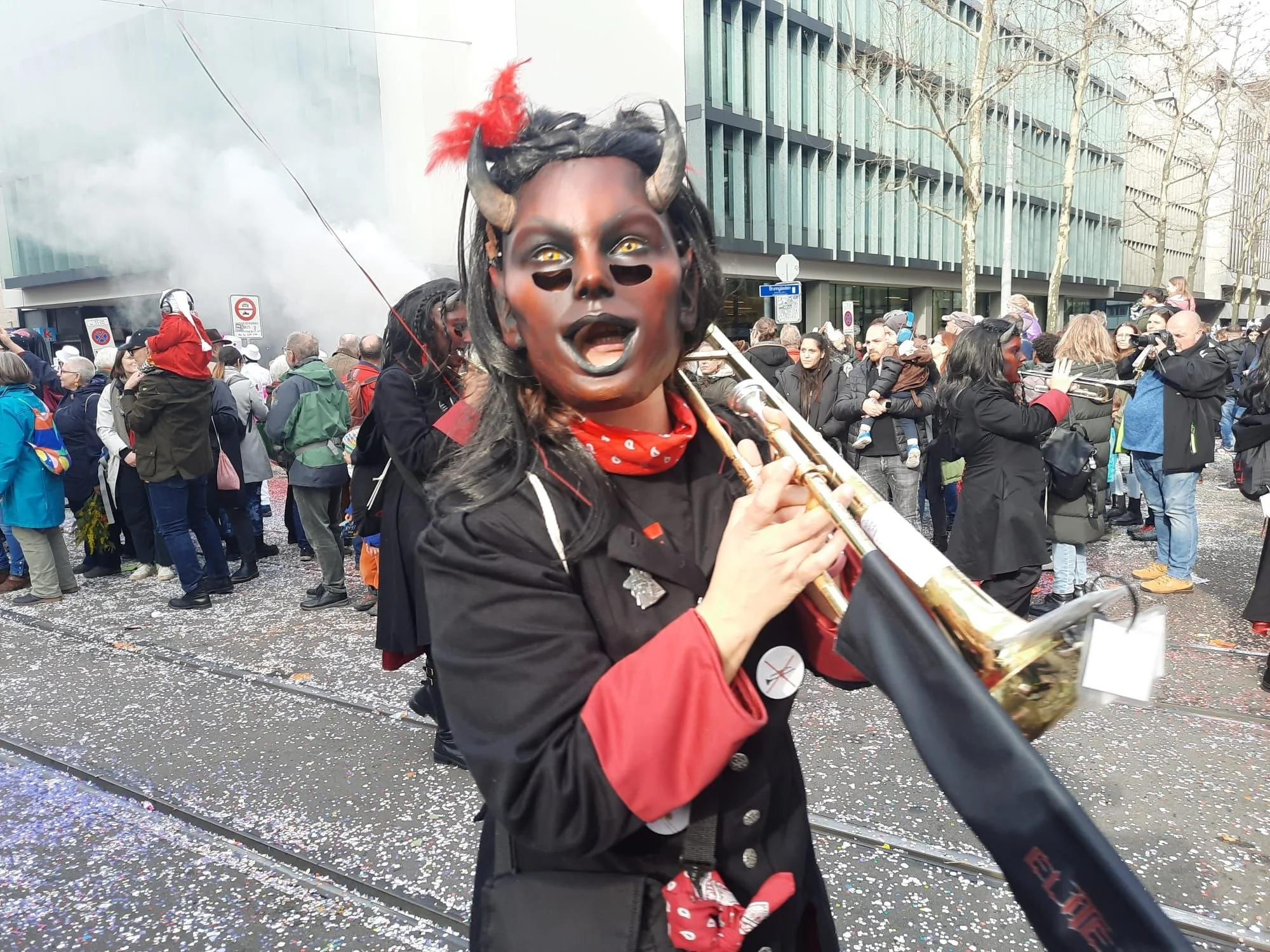 Eine Person trägt eine Teufelsmaske, Hörner, dunkle Hautfarbe und spielt Saxophon während eines Straßenfestivals oder einer Parade, umgeben von Menschen mit Bäumen und Gebäuden im Hintergrund.