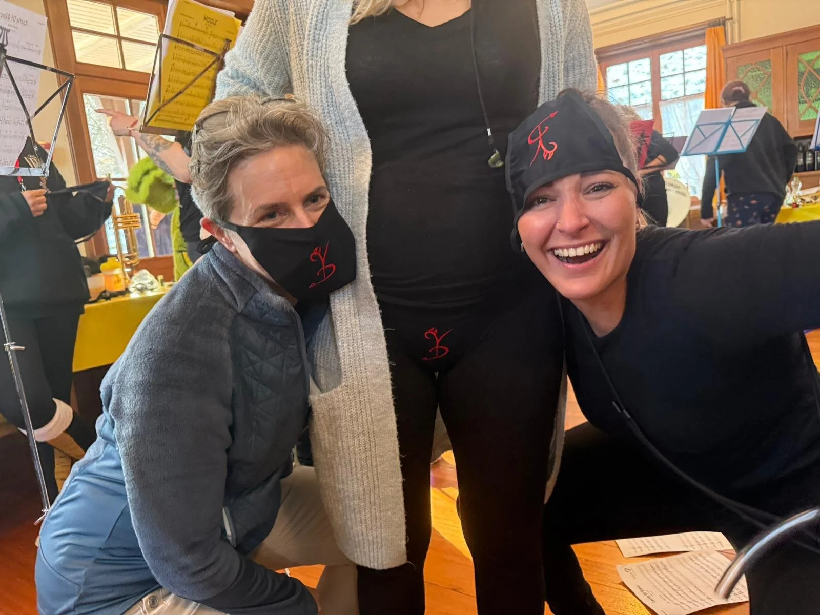 Drei Frauen, zwei tragen Schutzmasken, alle lachen und posieren für die Kamera in einem Raum mit großen Fenstern und Musiknotenständern im Hintergrund.
