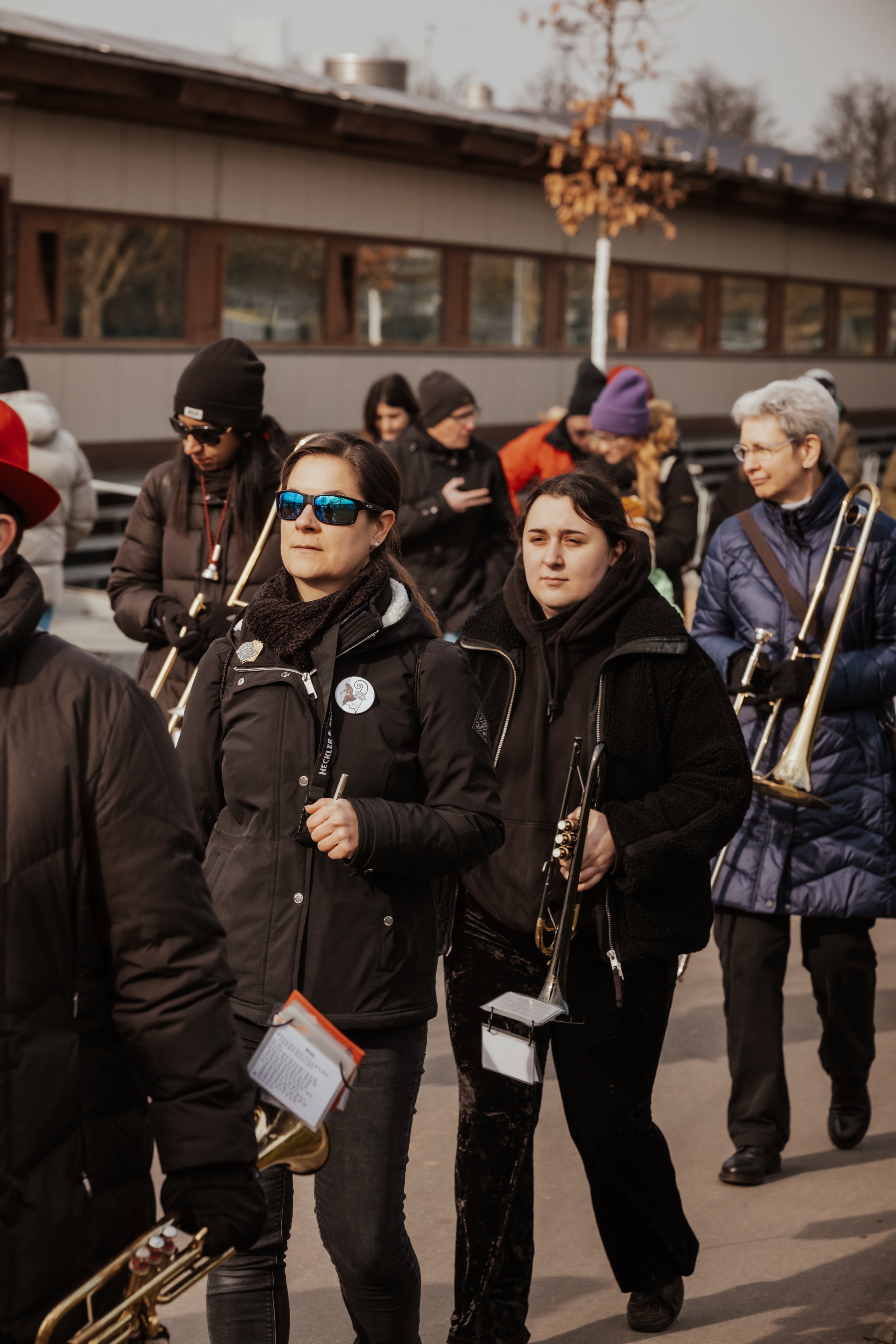 Gruppe von Menschen, die eine Marsch- oder Musikkapelle mit Blechblasinstrumenten bei einem Event im Freien begleiten, einige tragen Winterkleidung und Sonnenbrillen.