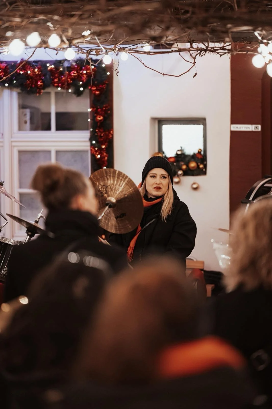Eine Frau mit schwarzen Haaren, eine schwarze Mütze und einem schwarzen Mantel sitzt in einem festlich dekorierten Raum, umgeben von mehreren Personen, vermutlich bei einem Weihnachts- oder Winterevent, mit Weihnachtsdekoration und Lichter im Hinterg