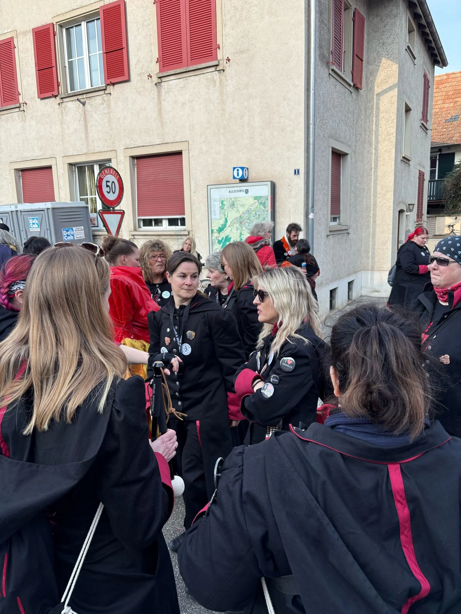 Gruppe von Menschen in dunklen Jacken, einige mit Pins und Buttons, stehen zusammen in einer Straße vor einem Gebäude mit roten Fensterläden, einige tragen rote Halstücher, es sieht aus wie eine Versammlung oder ein Treffen.