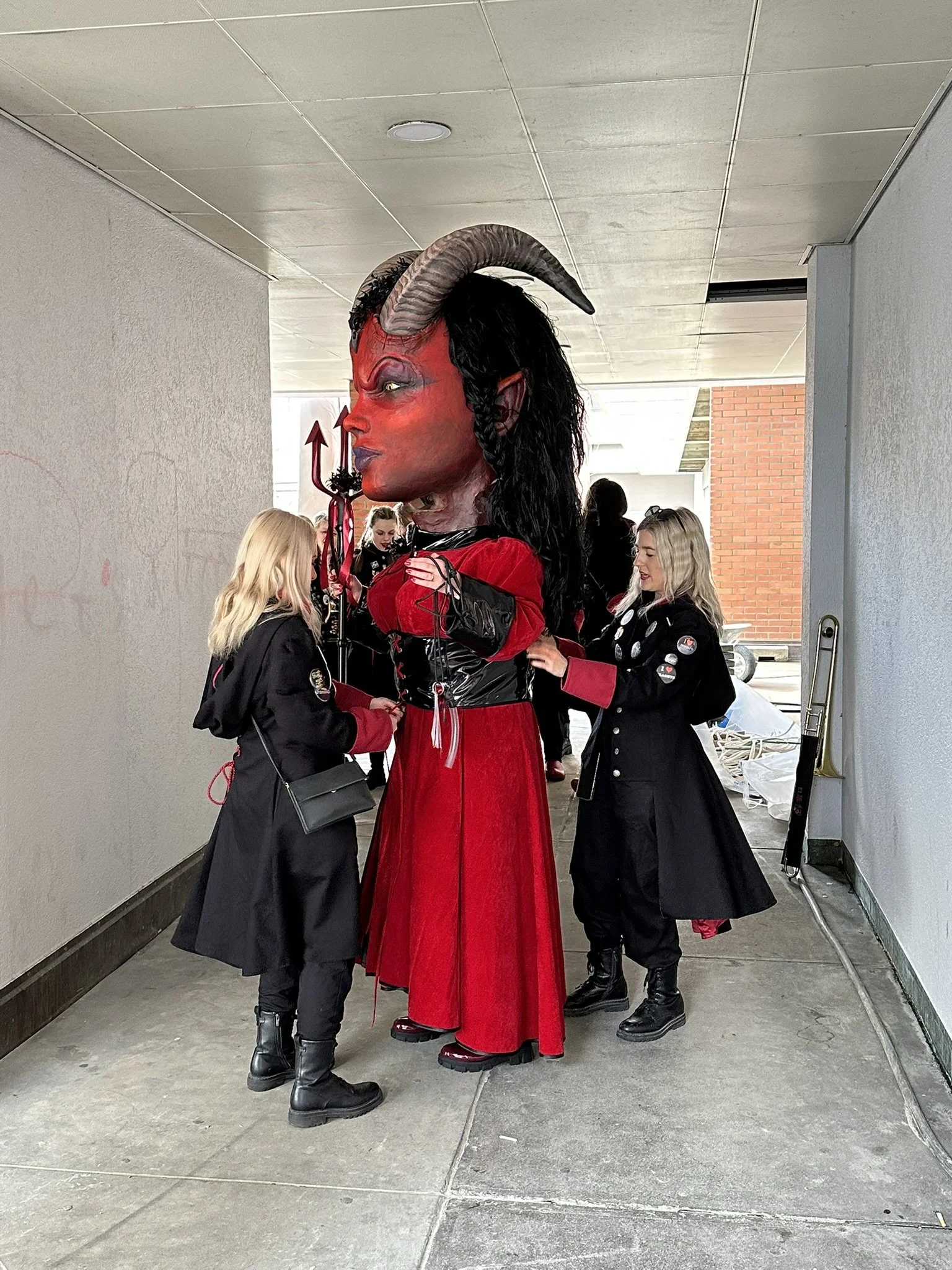 Damen sprechen mit einer großen Dämonen-Figur, die Hörner trägt, rote Kleidung und eine rote Hautfarbe hat, in einem dunklen Tunnel.