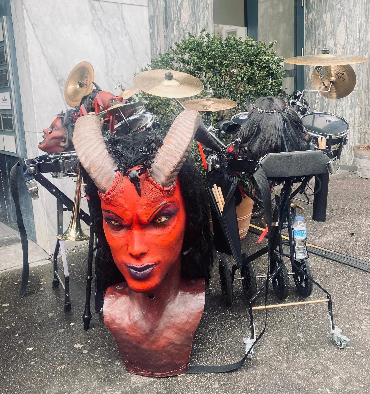 Eine Straßenkünstlerdekoration bestehend aus einer aufwändigen Teufelsmaske mit roten Gesicht und schwarzen Haaren, umgeben von Musikinstrumenten und Rollstühlen, im Hintergrund eine Pflanze und eine Wand mit Marmoroptik.