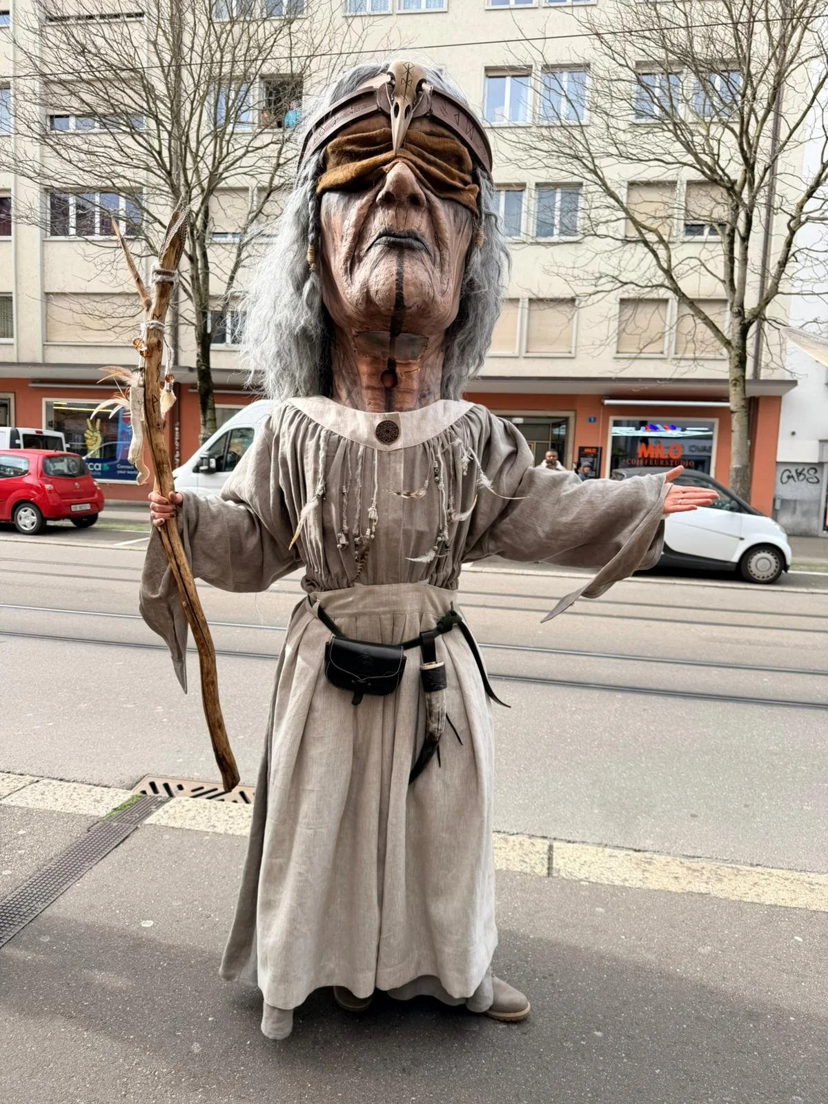 Eine Person in einem großen, aufwendig gestalteten Maskenkostüm als eine alte, weise Figur mit grauem Haar, Brille und einer langen Robe. Die Person hält einen Stock in der Hand und steht an einer Straße.