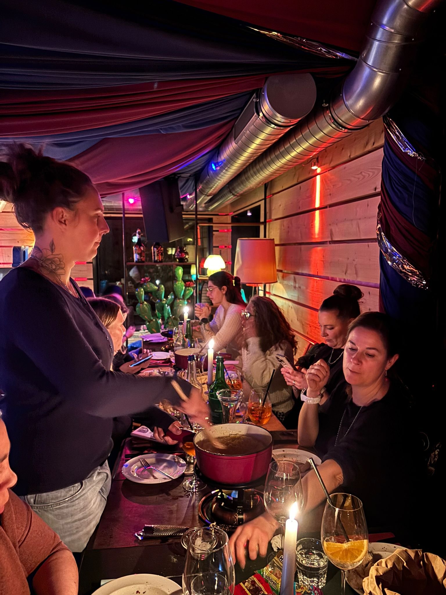 Eine Gruppe von Menschen sitzt an einem langen Tisch in einem Restaurant mit bunten Lichter und Dekorationen. Es gibt Getränke, Kerzen und einen Topf auf dem Tisch. Eine Frau steht und serviert Essen.