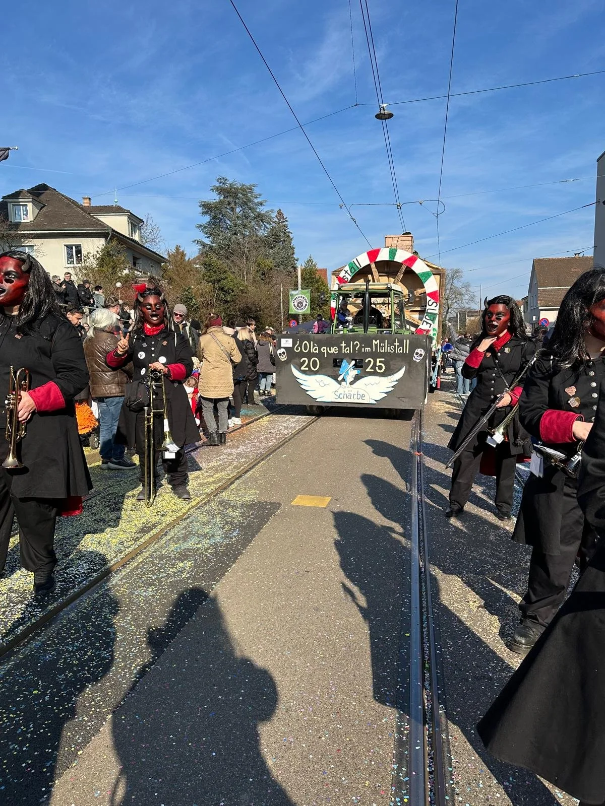 Menschen in Karnevalskostümen auf der Straße während eines Umzugs, mit einer Karnevalswagen im Hintergrund, bei schönem Wetter.