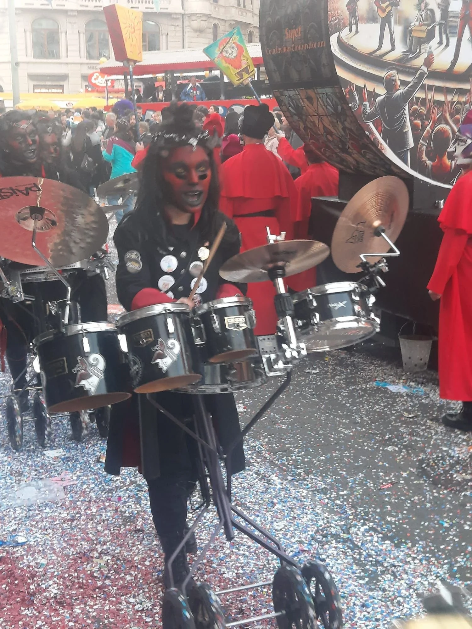 Person in Kostüm spielt Schlagzeug auf einer Feier, umgeben von Menschen im roten Outfit und bunten Verkleidungen, mit Konfetti auf dem Boden.