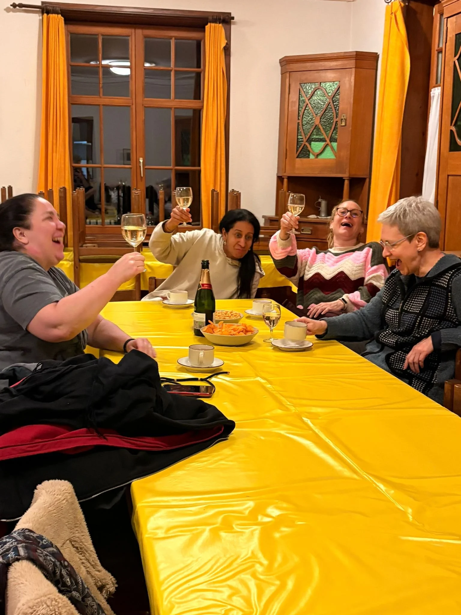 Vier Frauen sitzen um einen Tisch, lachen und stoßen mit Weingläsern an, während sie Getränke trinken. Der Tisch ist mit einer gelben Tischdecke bedeckt und es gibt Snacks. Im Hintergrund sind gelbe Vorhänge und eine Holzwand.