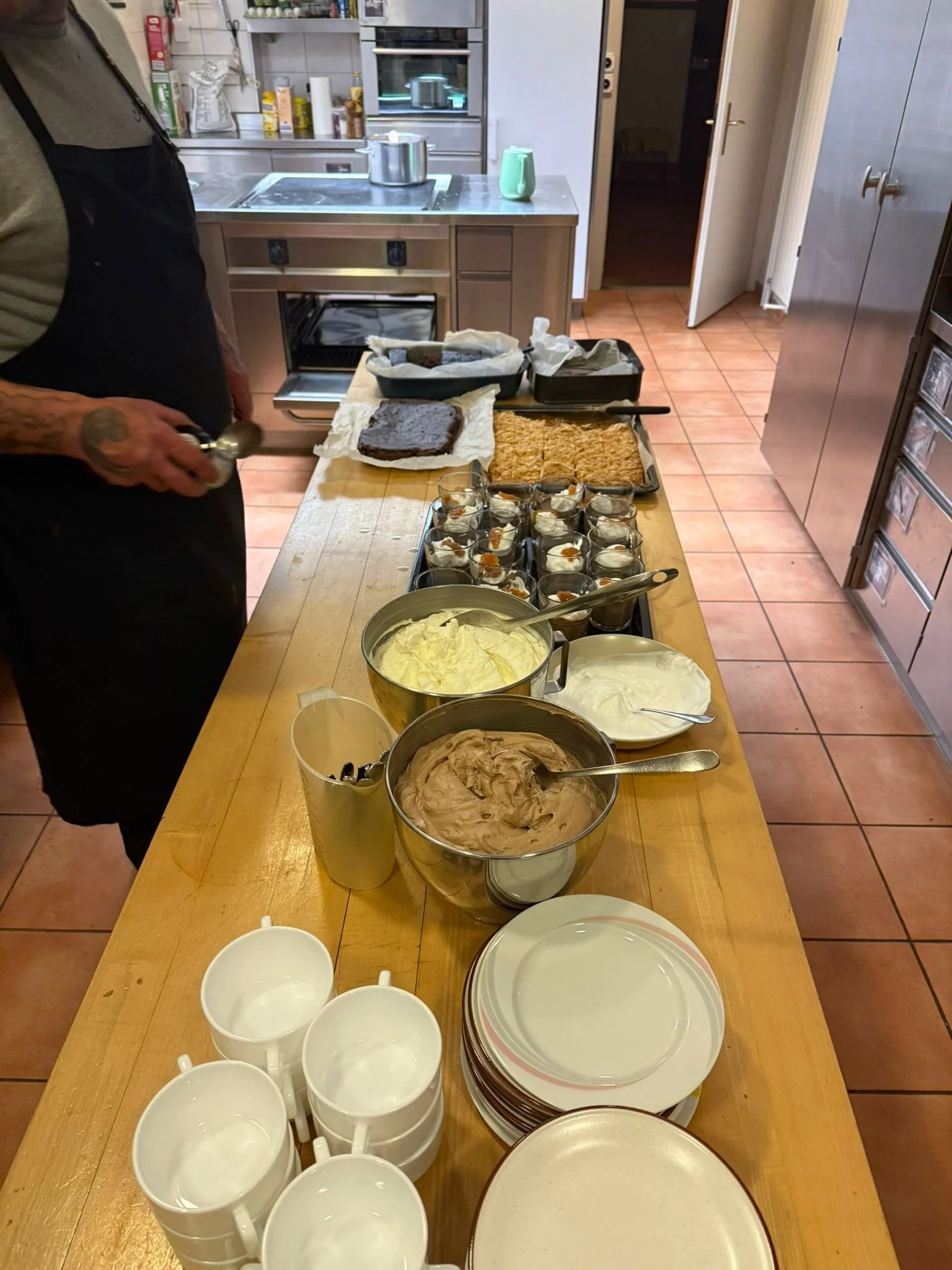 Büfetttisch mit mehreren Schüsseln mit Eiscreme, Tassen, Tellern, und auf dem Tisch sind auch Torten und kleine Gläser mit Desserts in einem Küchenraum.