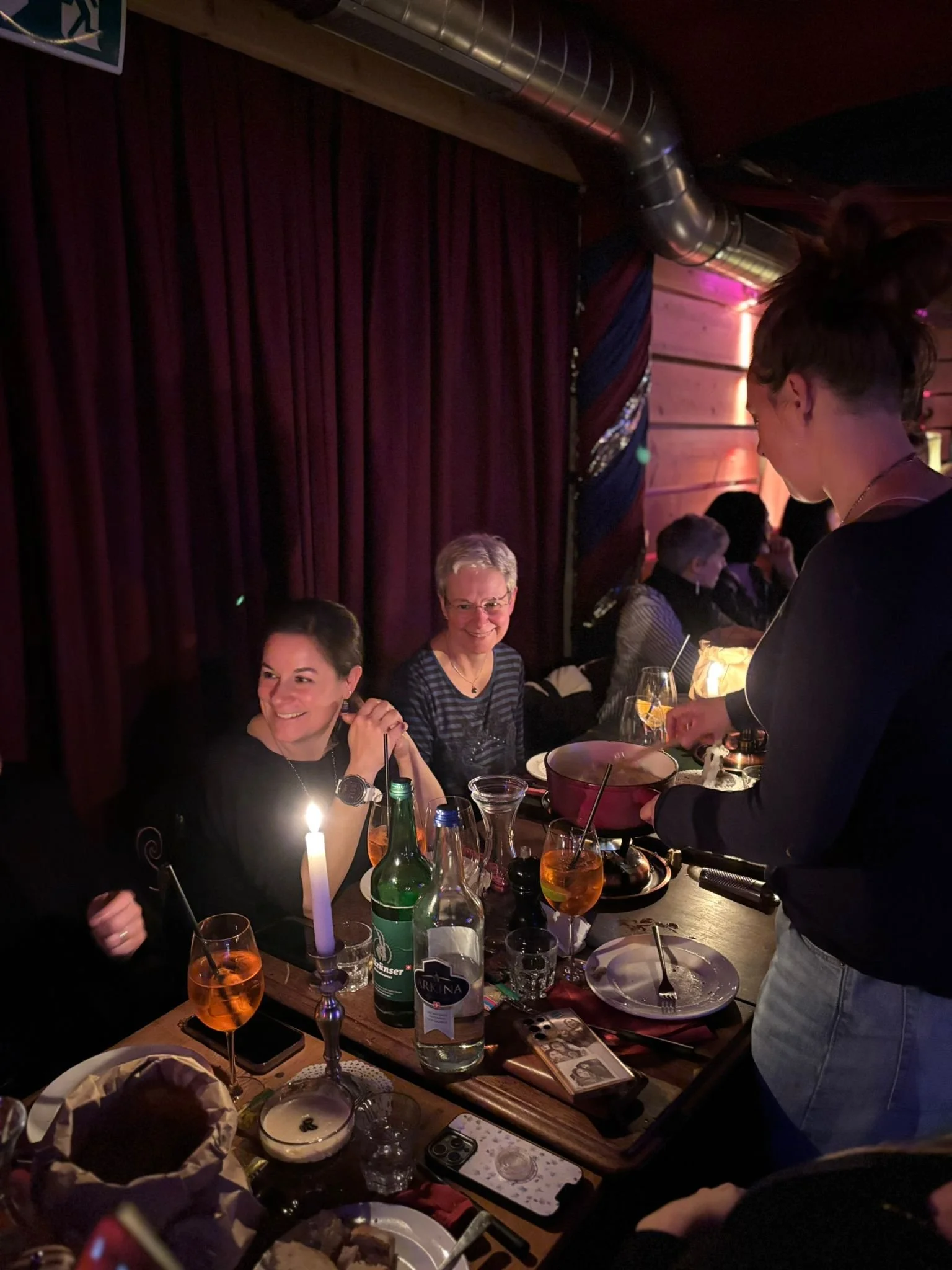 Menschen sitzen an einem Tisch in einem dunklen Raum, eine Frau wird bedient, es gibt Getränke und Kerzen auf dem Tisch.