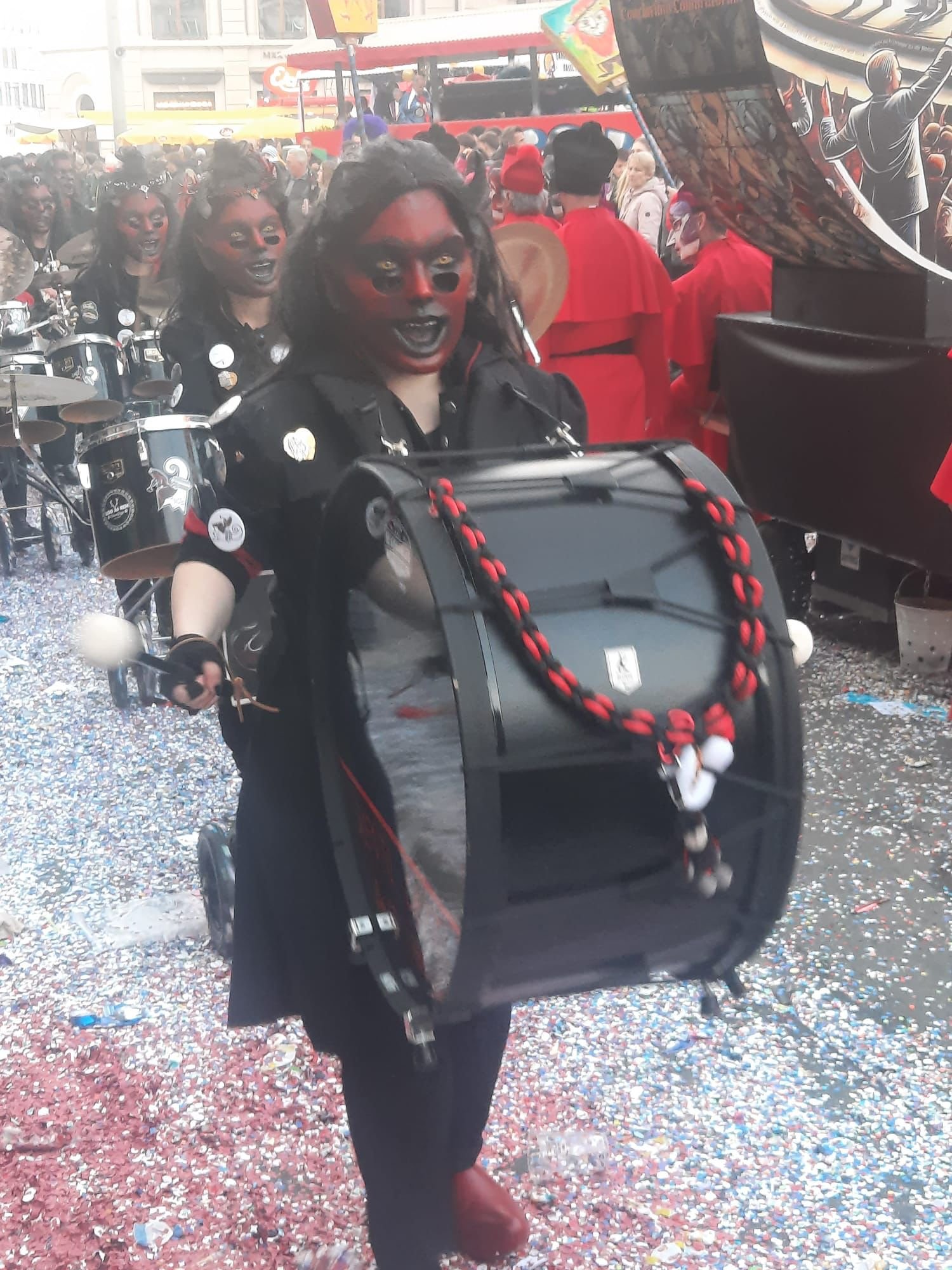 Menschen in fantasievollem Kostüm mit schwarzer Haut und roten Gesichtern bei einer Parade, umgeben von Konfetti, auf einem geschäftigen Stadtfest