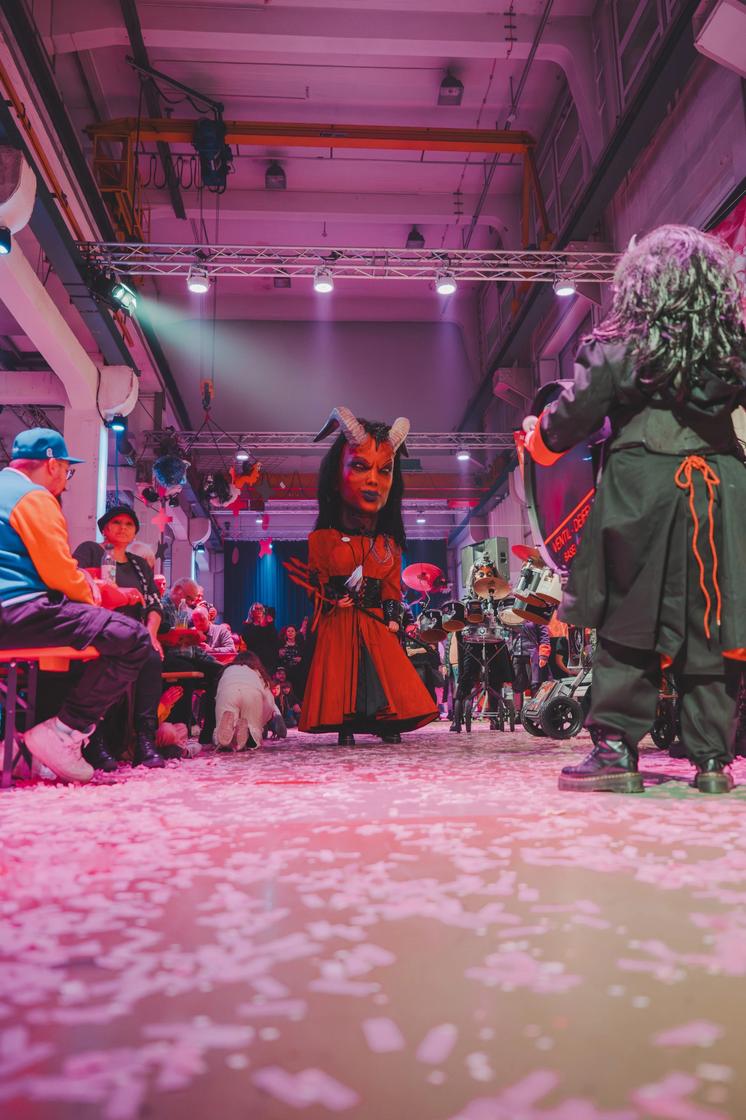 Mensch in Teufelskostüm auf Laufsteg bei Karnevalsveranstaltung, mit Publikum im Hintergrund, rosa und blaue Beleuchtung, Konfetti auf Boden.