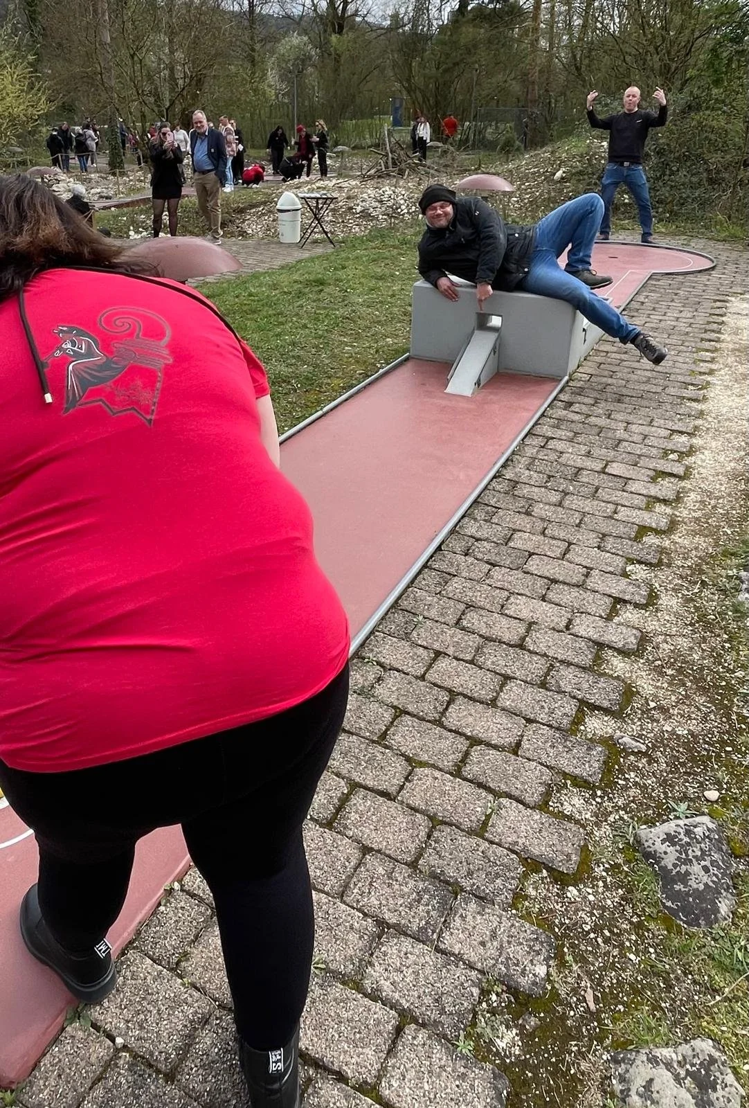 Menschen spielen Minigolf im Freien auf einer gepflegten Anlage mit Bäumen im Hintergrund, einige Zuschauer beobachten die Aktivität.