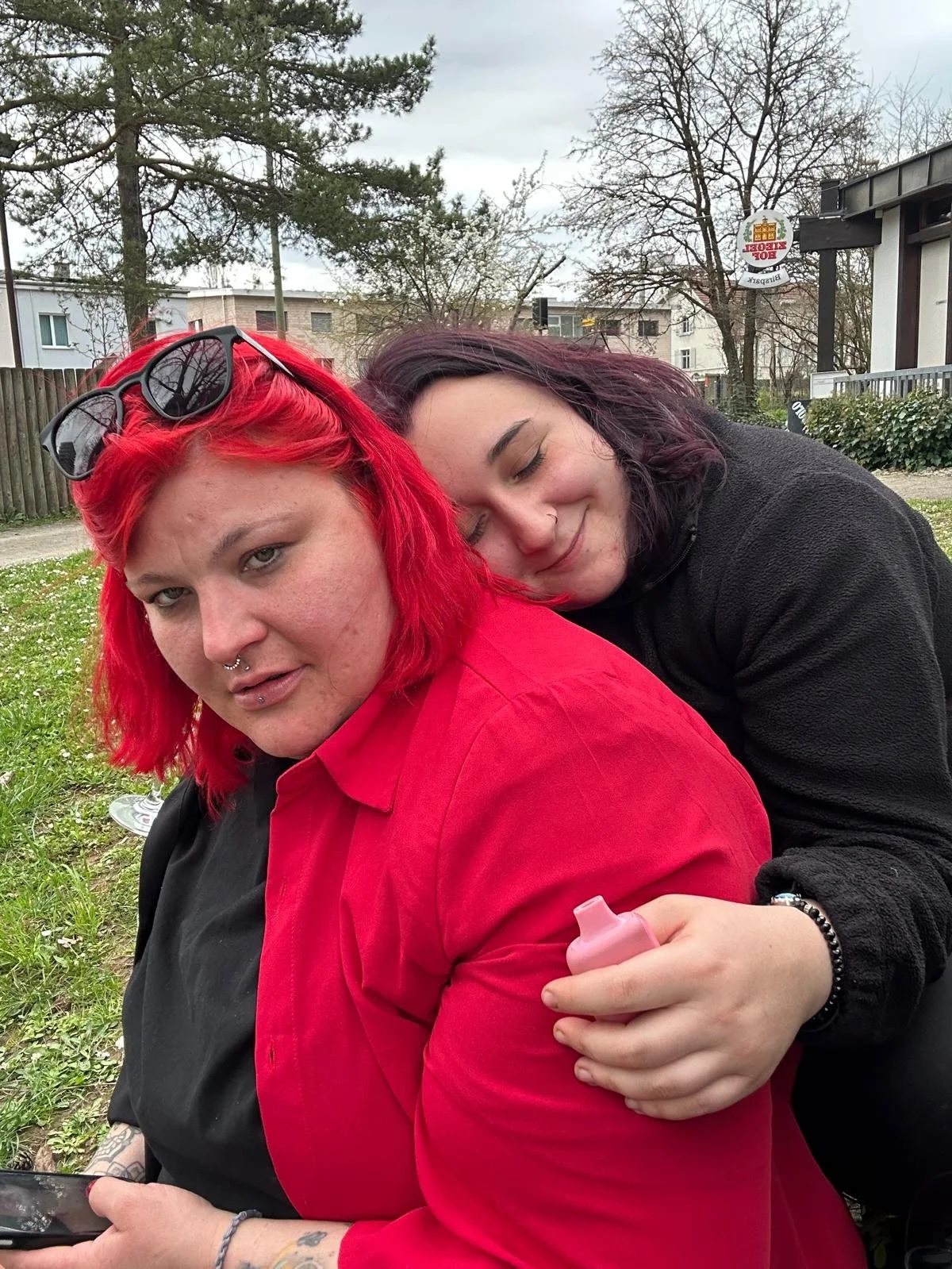 Zwei Frauen sitzen im Freien, die eine hat leuchtend rote Haare und trägt eine rote Jacke, die andere hat dunkle Haare und trägt eine schwarze Jacke, sie umarmen sich freundlich.