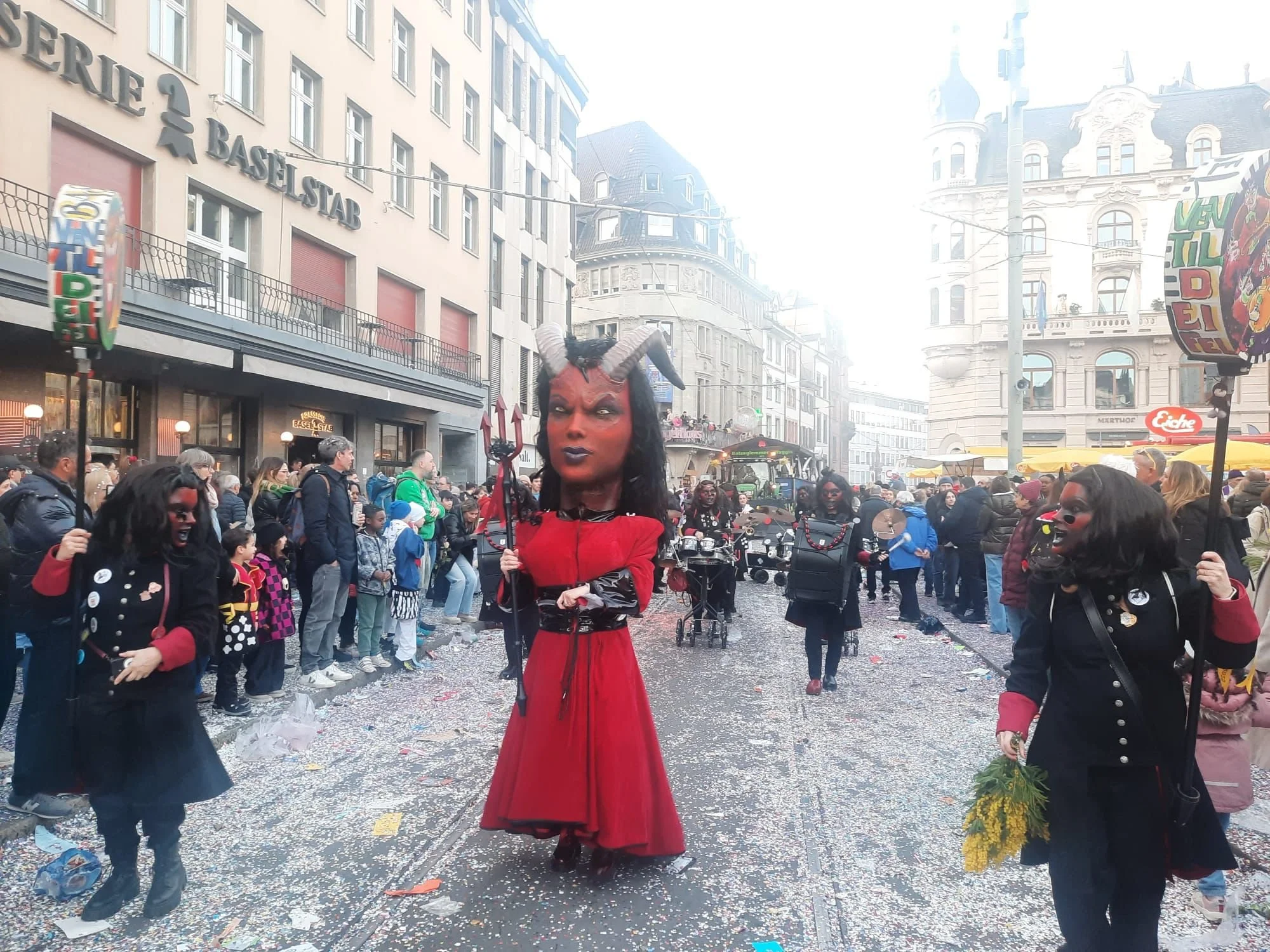 Menschen in Karnevals- oder Faschkostümen, darunter eine Frau mit Teufel-Perücke und Hornmasken, die an einer Straßenparade teilnehmen, mit Gebäuden im Hintergrund.