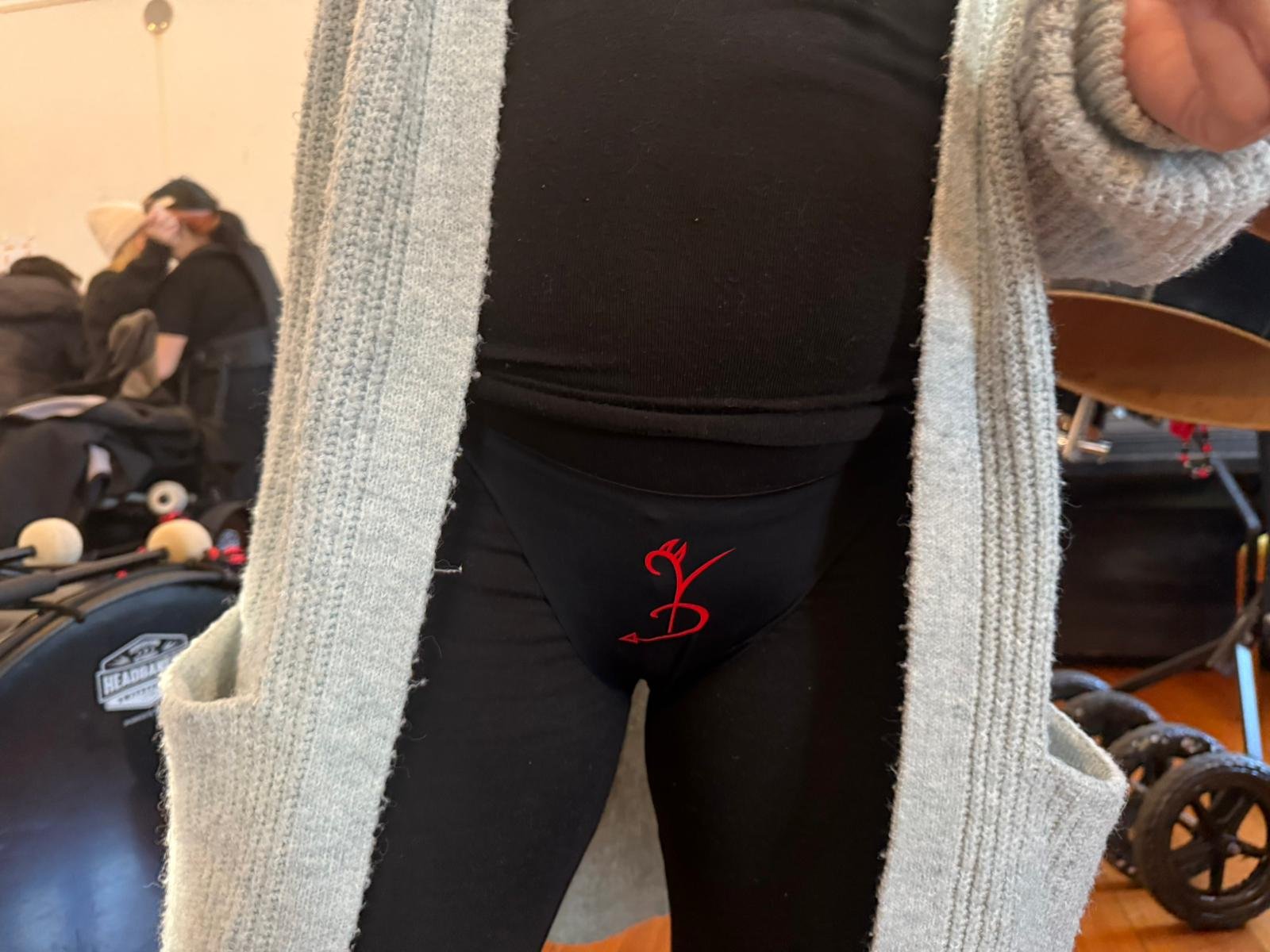 Nahaufnahme einer Person in einem schwarzen Outfit, die Unterwäsche mit rotem Logo trägt, umgeben von Kinderwagen und anderen Gegenständen im Hintergrund.
