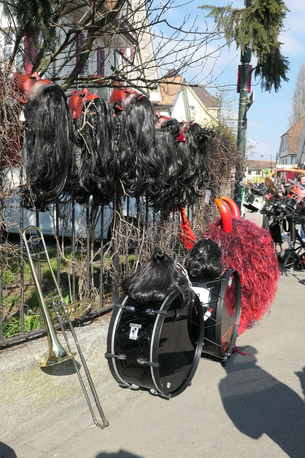 Masken, die wie schwarze und rote Haare und Hörner aussehen, hängen an einem Zaun. Es gibt auch einen Schlagzeug und eine große Trommel, die jedoch nicht getragen werden. In der Nähe steht eine Posaune. Die Szene ist auf einer Straße bei schönem Wett