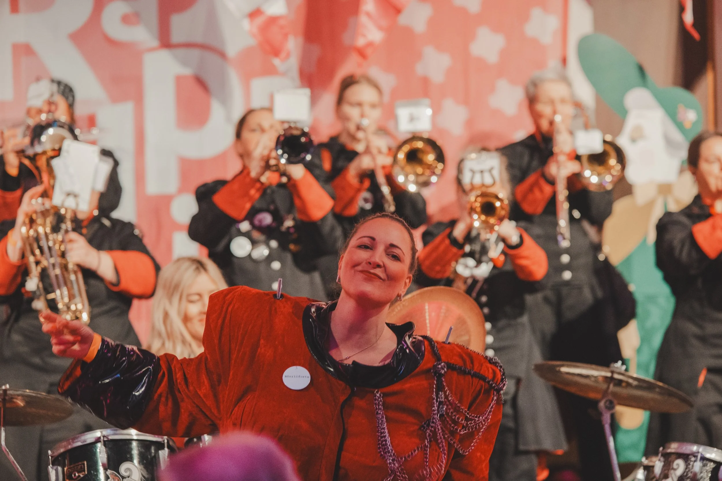 Eine Frau in roter Kleidung, die bei einer musikalischen Darbietung vor einer Gruppe von spielenden Musikerinnen und Musikern mit Trompeten und Saxophonen posiert.