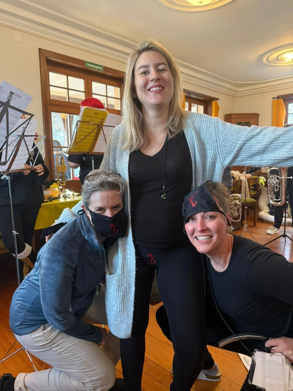 Drei Frauen in einem Raum, eine steht in der Mitte und die anderen beiden sind unten links und rechts. Sie scheinen bei einer Veranstaltung oder Probe mit Musikinstrumenten im Hintergrund zu sein.