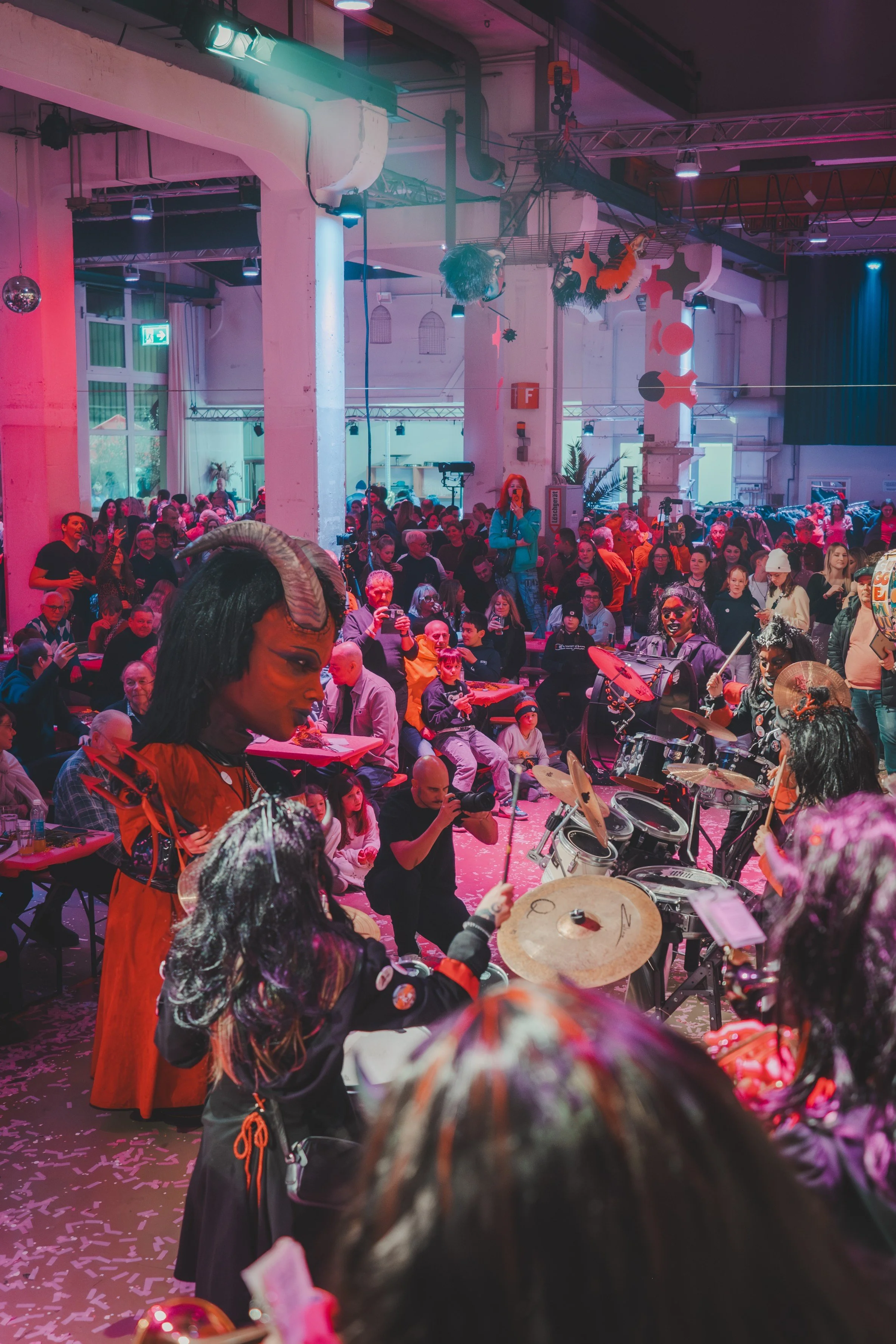 Eine große Gruppe Menschen bei einer Veranstaltung in einem Industriehallen-ähnlichen Raum mit bunten Lichtern. Im Vordergrund spielen Menschen in Kostümen, darunter eine mit Hörnern und eine mit einer schwarzen Perücke, während ein Schlagzeug gespie