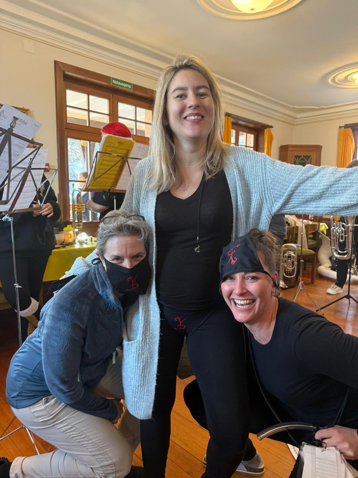 Drei Frauen, eine trägt eine graue Jacke, eine andere schwarze Kleidung, stehen und sitzen im Raum mit einem Blasinstrument in der Hand, im Hintergrund ein Musikensemble mit Notenständern und Instrumenten