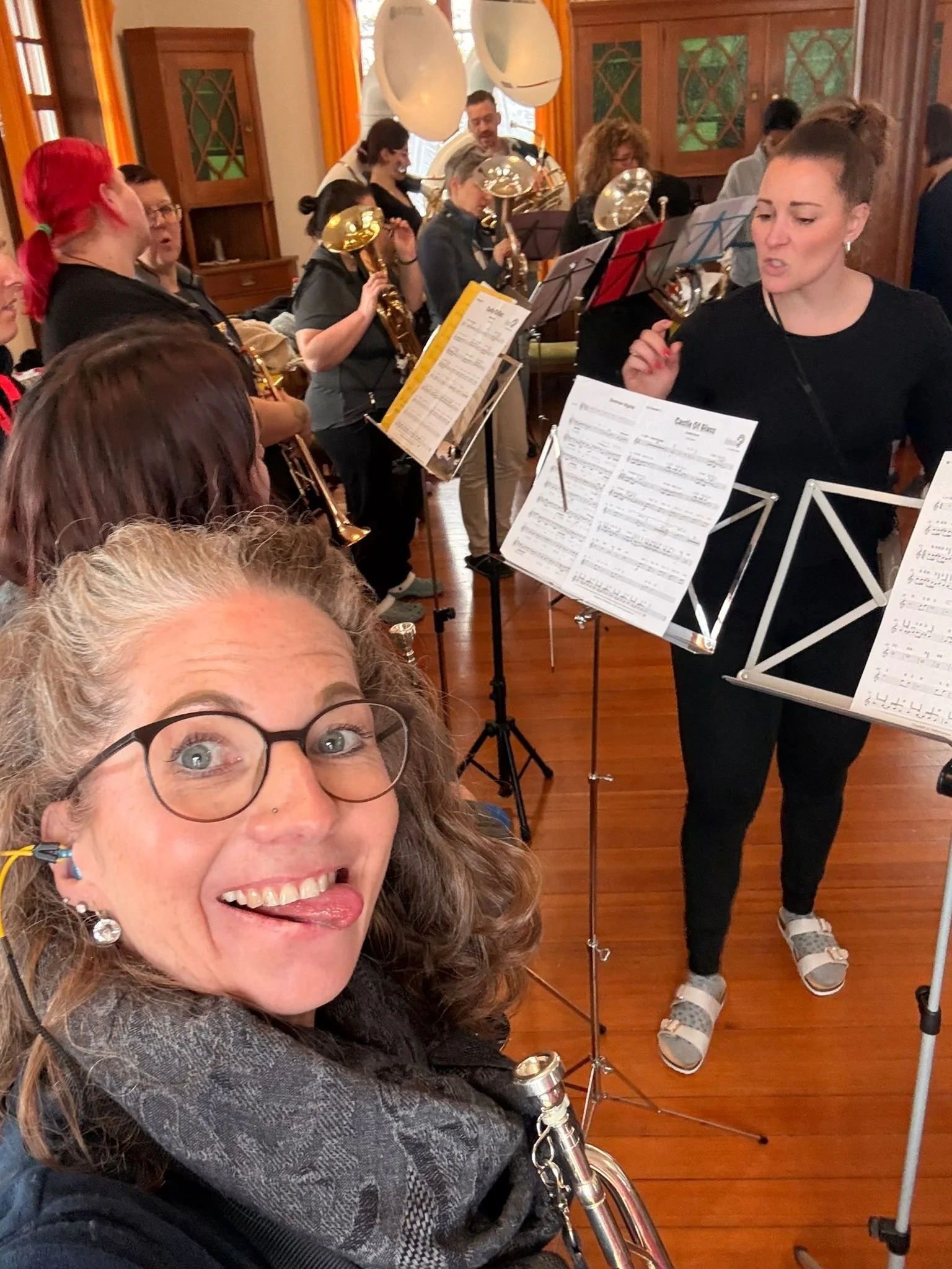 Eine Frau mit Brille und Locken, die einen Saxophon um den Hals trägt, macht ein Selfie vor einem Orchester in einem Holzraum, während Musiker mit Instrumenten proben.