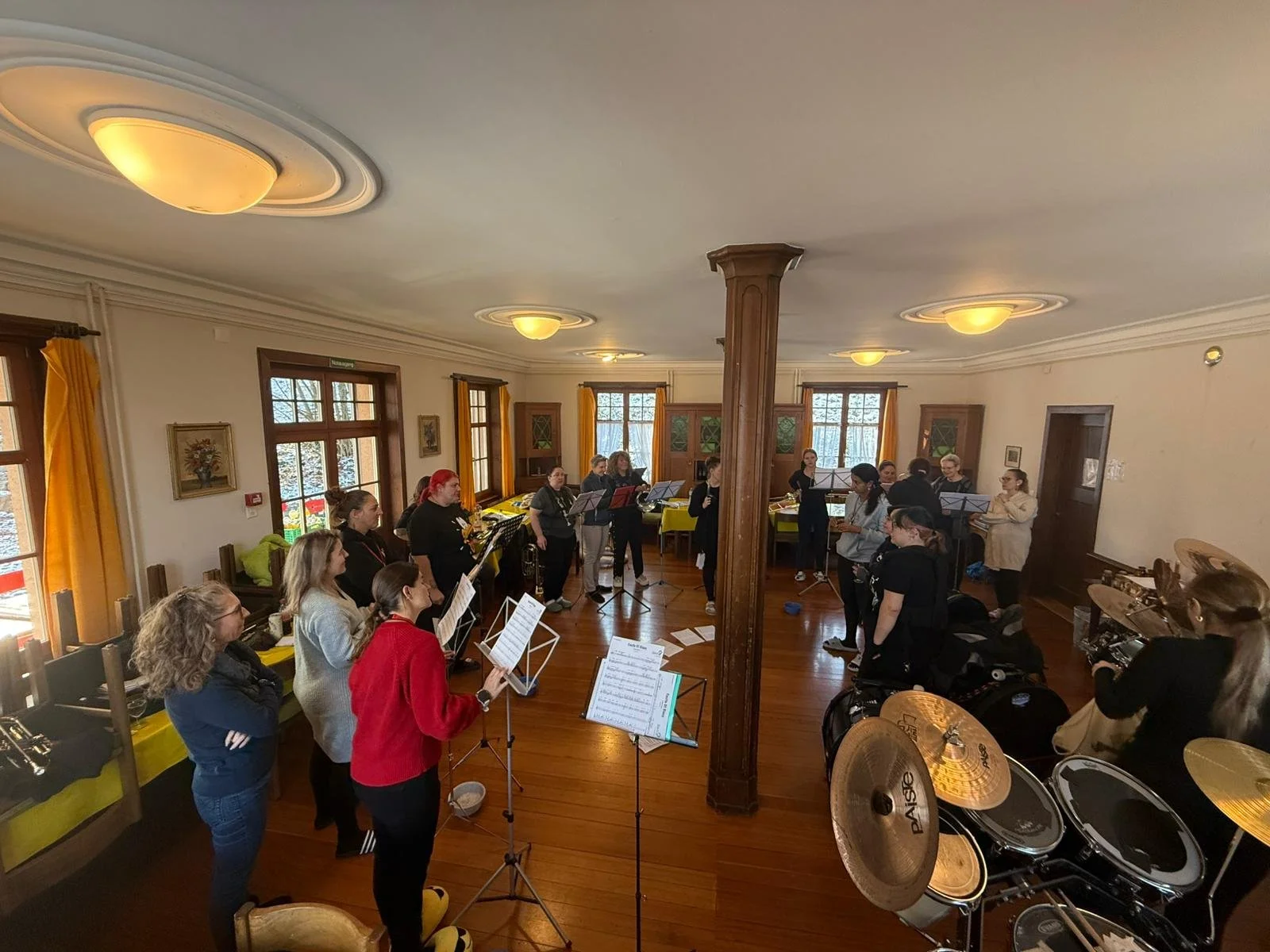 Gruppe von Frauen, die in einem geräumigen, hell erleuchteten Wohnzimmer mit Holzboden und großen Fenstern proben, umgeben von Musikinstrumenten und Notenständern.