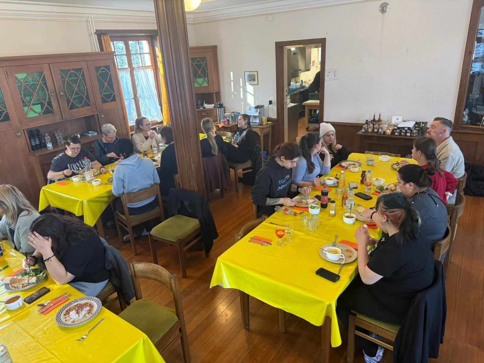 Mehrere Personen sitzen an Tischen mit gelben Tischdecken in einem Raum und essen.