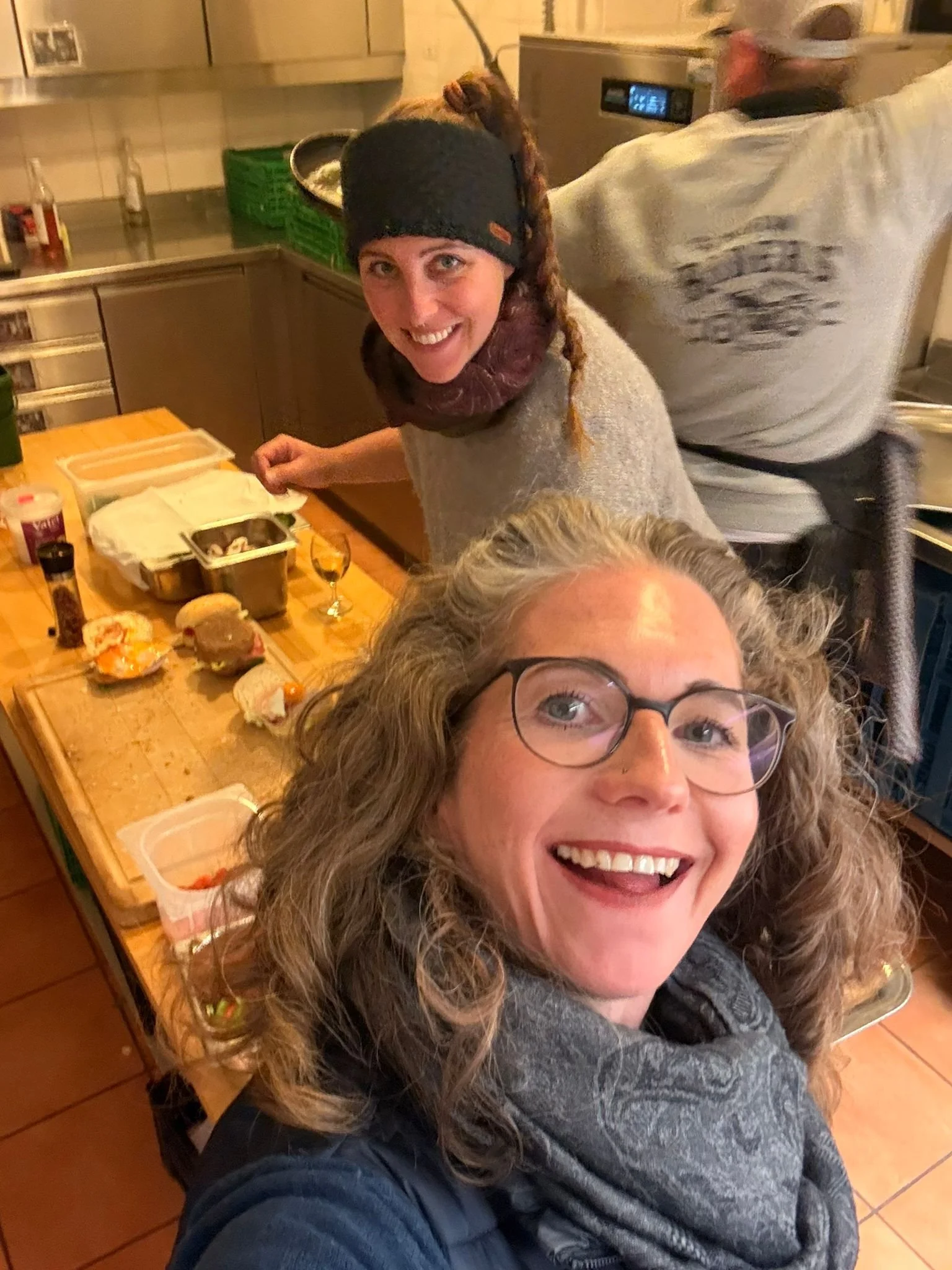 Zwei Frauen in einer Küche, eine lächelnd in die Kamera schauend, die andere beim Vorbereiten von Speisen, im Hintergrund ein Mann, der sich mit einem Küchengerät beschäftigt.