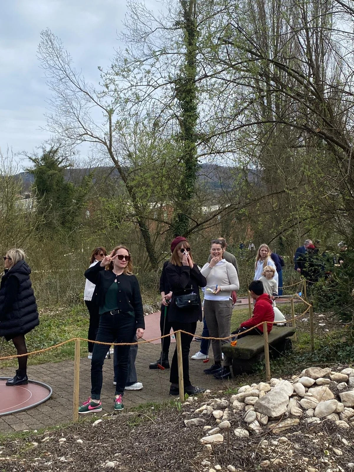 Grupp People im Park mit Bäumen und Natur, einige sitzen auf einer Bank, andere stehen und unterhalten sich, Kinder und Erwachsene, es ist bewölkter Himmel.