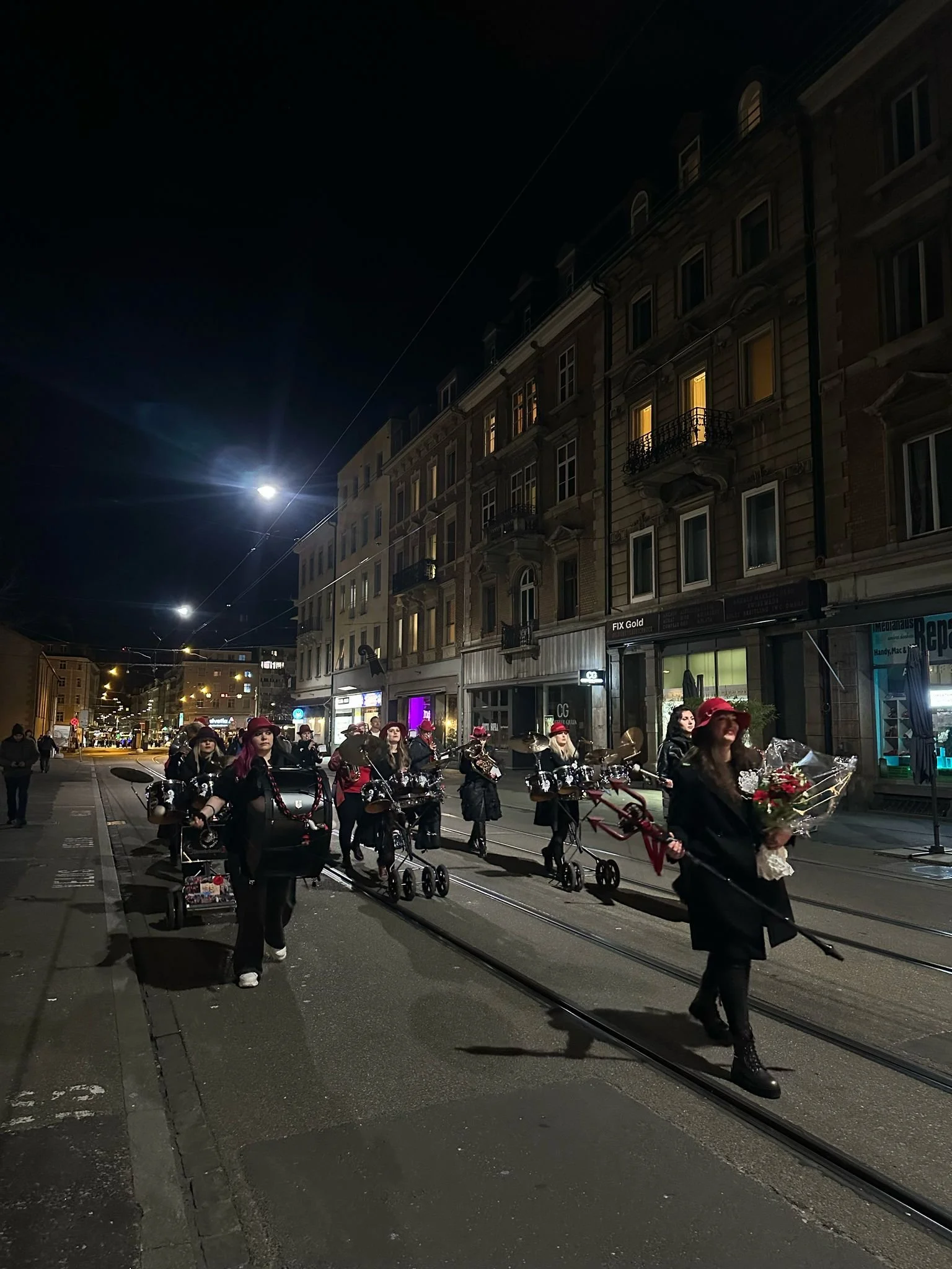 Frauen gehen nachts auf einer Straßenbahn-Trasse, eine von ihnen trägt einen Blumenstrauß und einen Regenschirm, alle sind in dunkler Kleidung und roten Hüten.