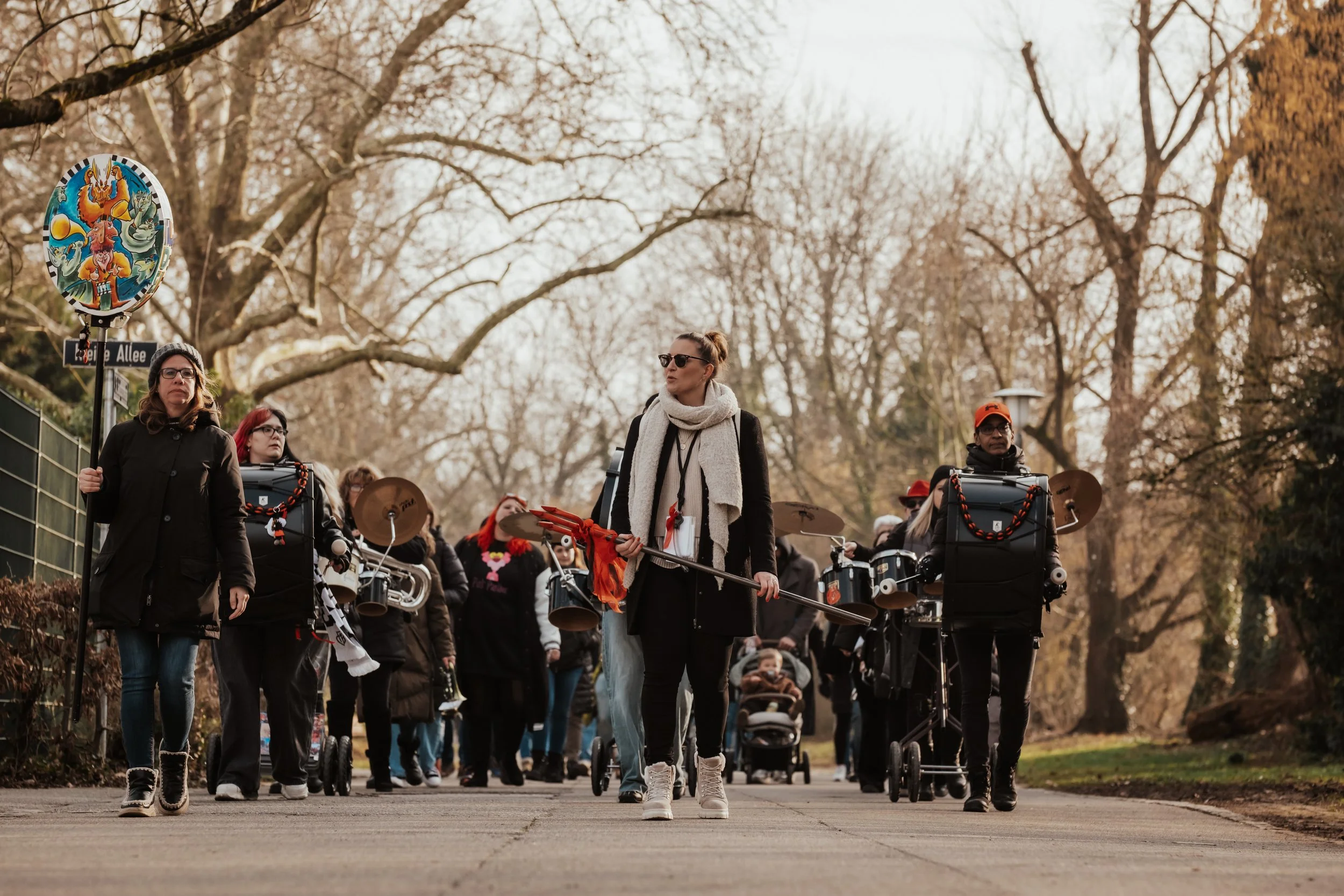 Eine Gruppe von Menschen läuft auf einem Weg im Park, einige tragen Musikinstrumente wie Trommeln und cymbals, während eine Frau in der Mitte ein Speer hält und Sonnenbrille trägt.