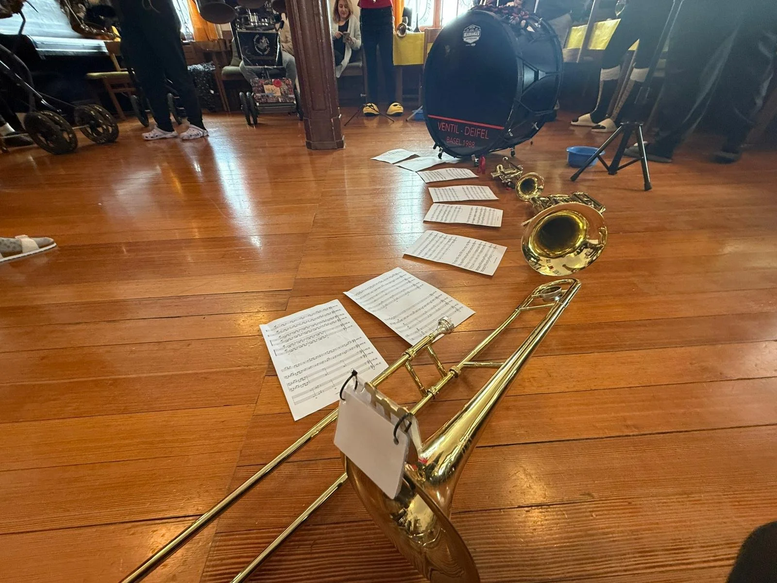 Ein auf dem Boden liegendes Saxophon, umgeben von gedruckten Notenblättern, in einem Raum mit Holzfußboden, im Hintergrund sind Menschen, Instrumentenkoffer und andere Musikinstrumente zu sehen.