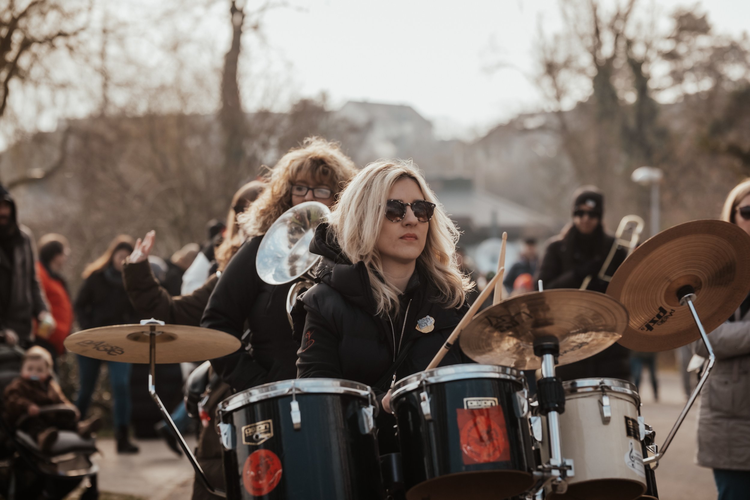 Junge Frau spielt auf einem Schlagzeug bei einer Demonstration im Freien, weitere Menschen im Hintergrund.