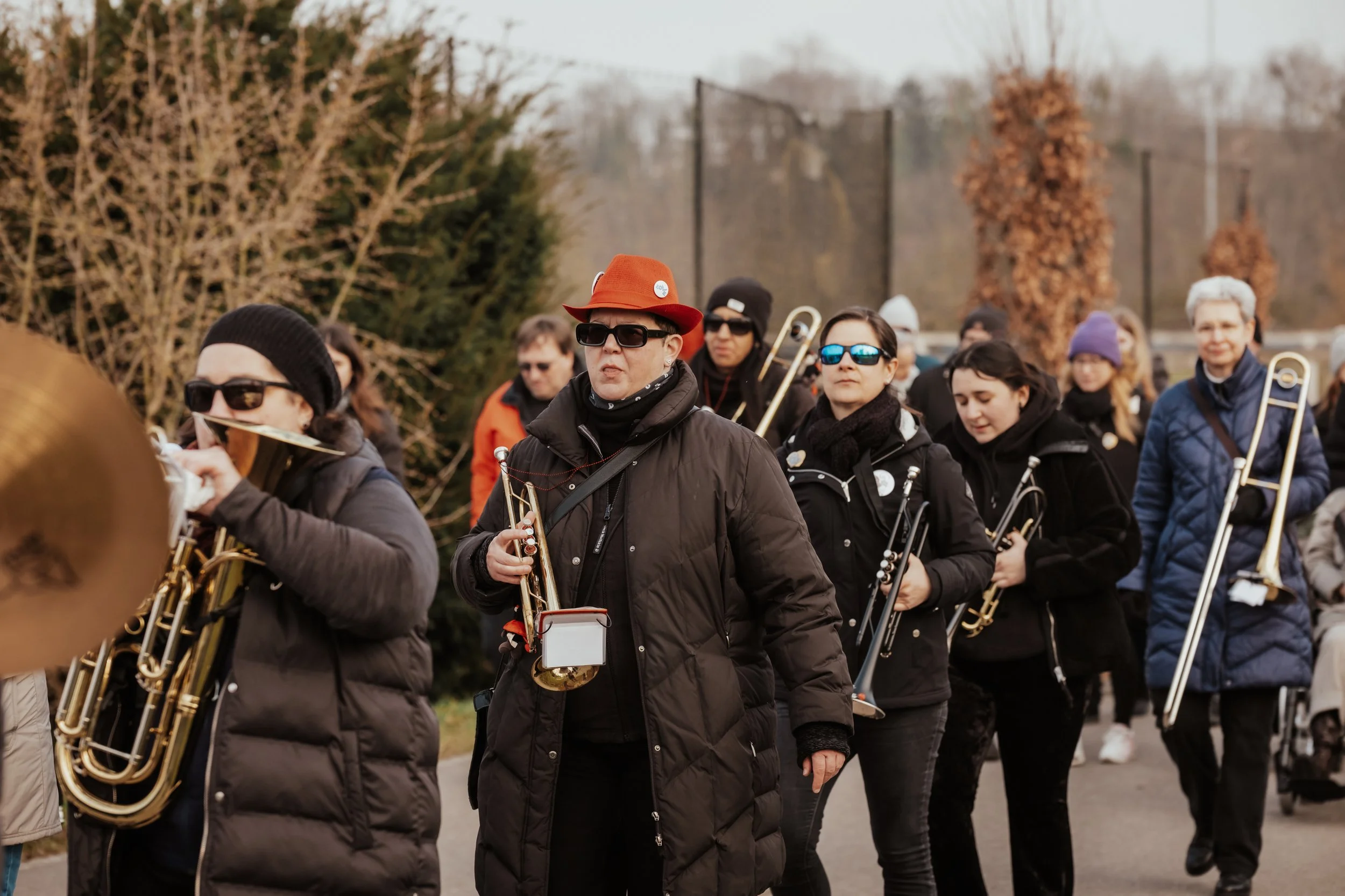 Eine Gruppe von Menschen, die Musikinstrumente wie Trompeten und Posaunen tragen, unterwegs im Freien bei kühlem Wetter, mit bunten Jacken und Sonnenbrillen, umgeben von Herbstbäumen.
