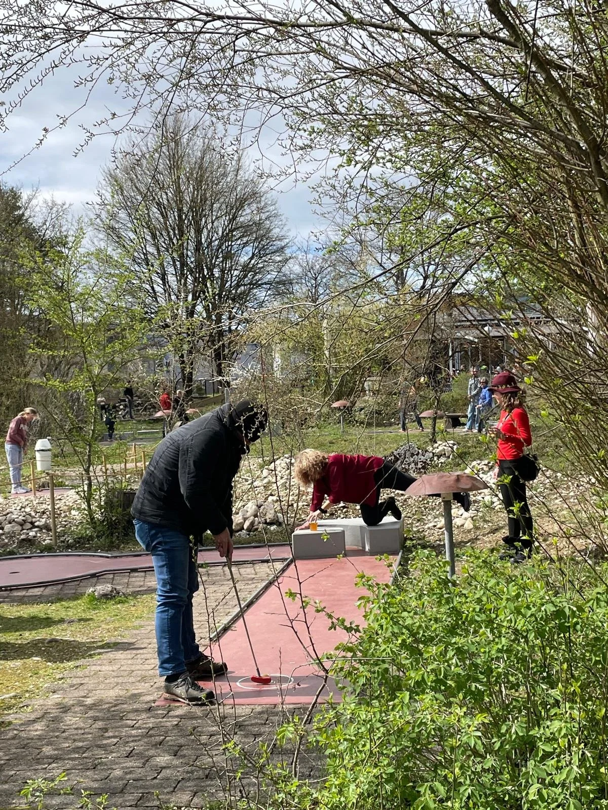 Menschen beim Mini-Golf spielen in einem Garten mit Bäumen und Büschen an einem bewölkten Tag.