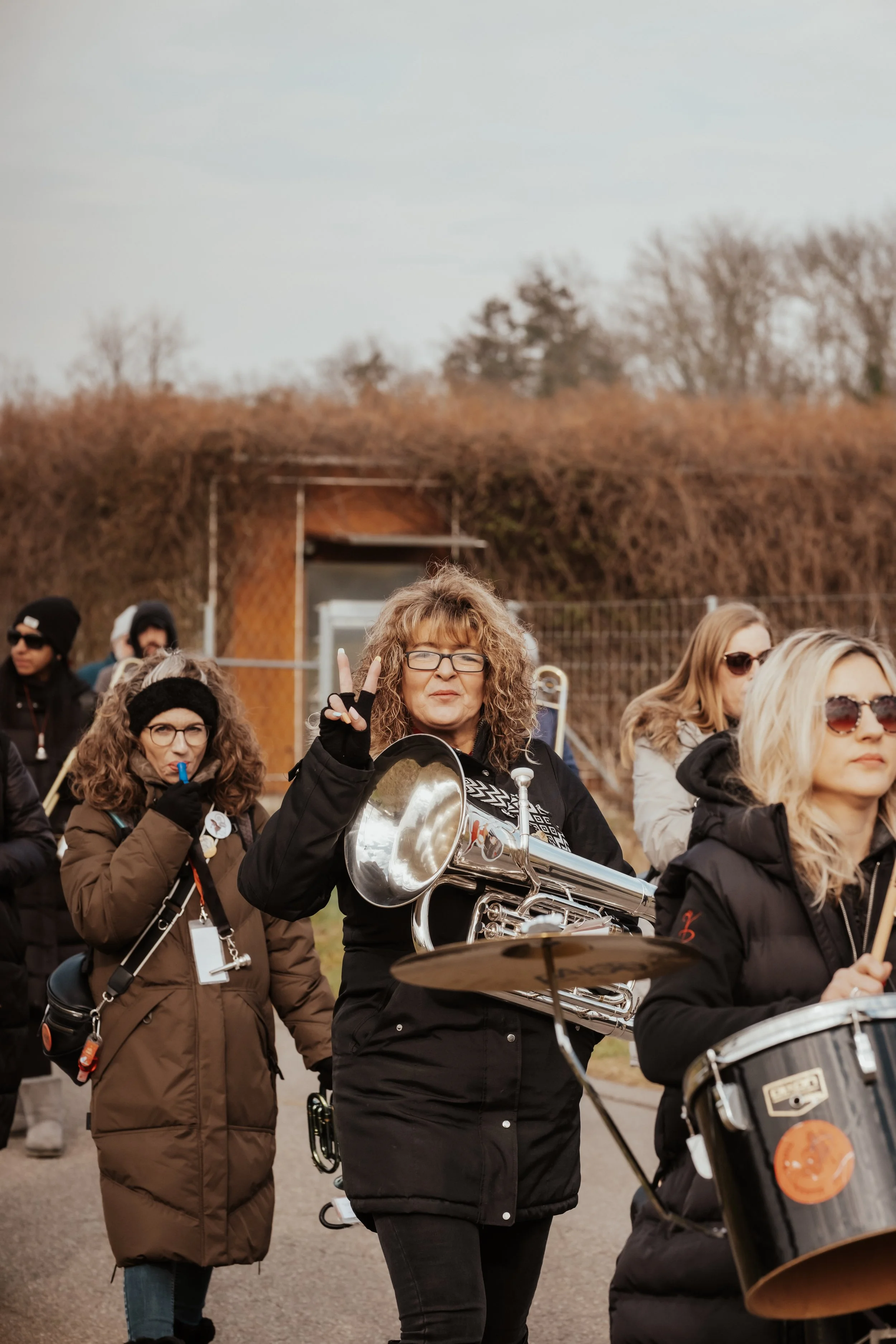 Eine Gruppe von Frauen mit Musikinstrumenten, eine Frau macht ein Peace-Zeichen und lächelt, im Hintergrund sind Bäume und ein Zaun bei kühlem Wetter.