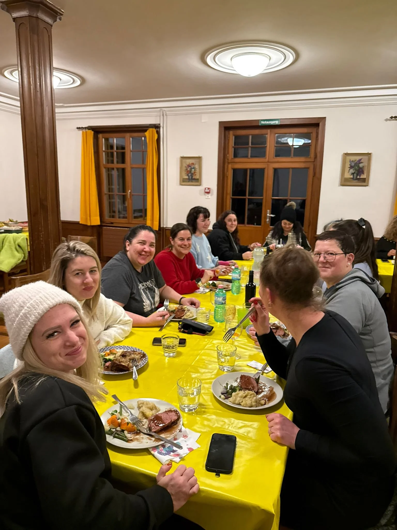 Gruppe von Menschen beim Essen an einem langen Tisch in einem Raum mit Holzwänden und gelben Vorhängen.
