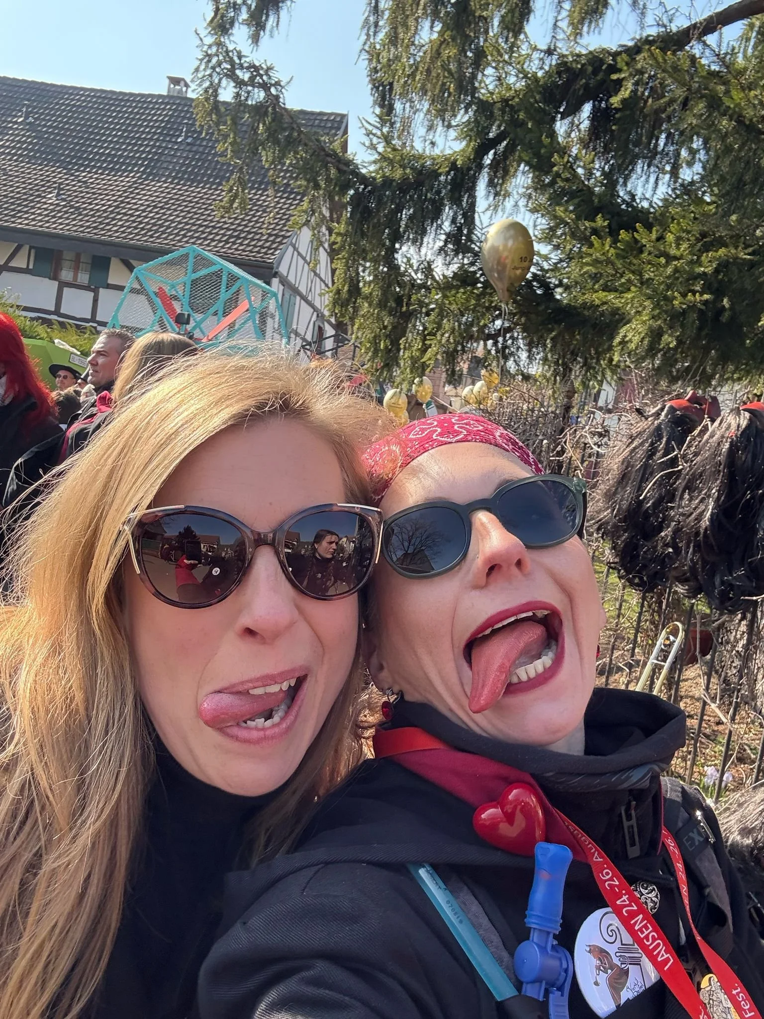 Zwei lachende Frauen mit Sonnenbrillen, die wild die Zunge herausstrecken, während eine im Freien bei einem Fest oder Festival posiert.